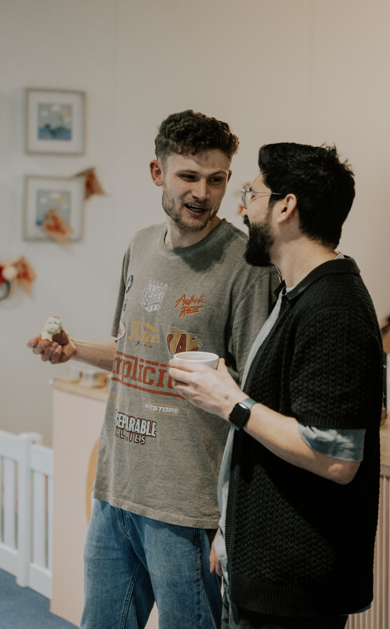 Two men are engaged in conversation at a social gathering. One is holding a cupcake, the other a cup. They are smiling and standing in a room with framed pictures and decorations on the wall.