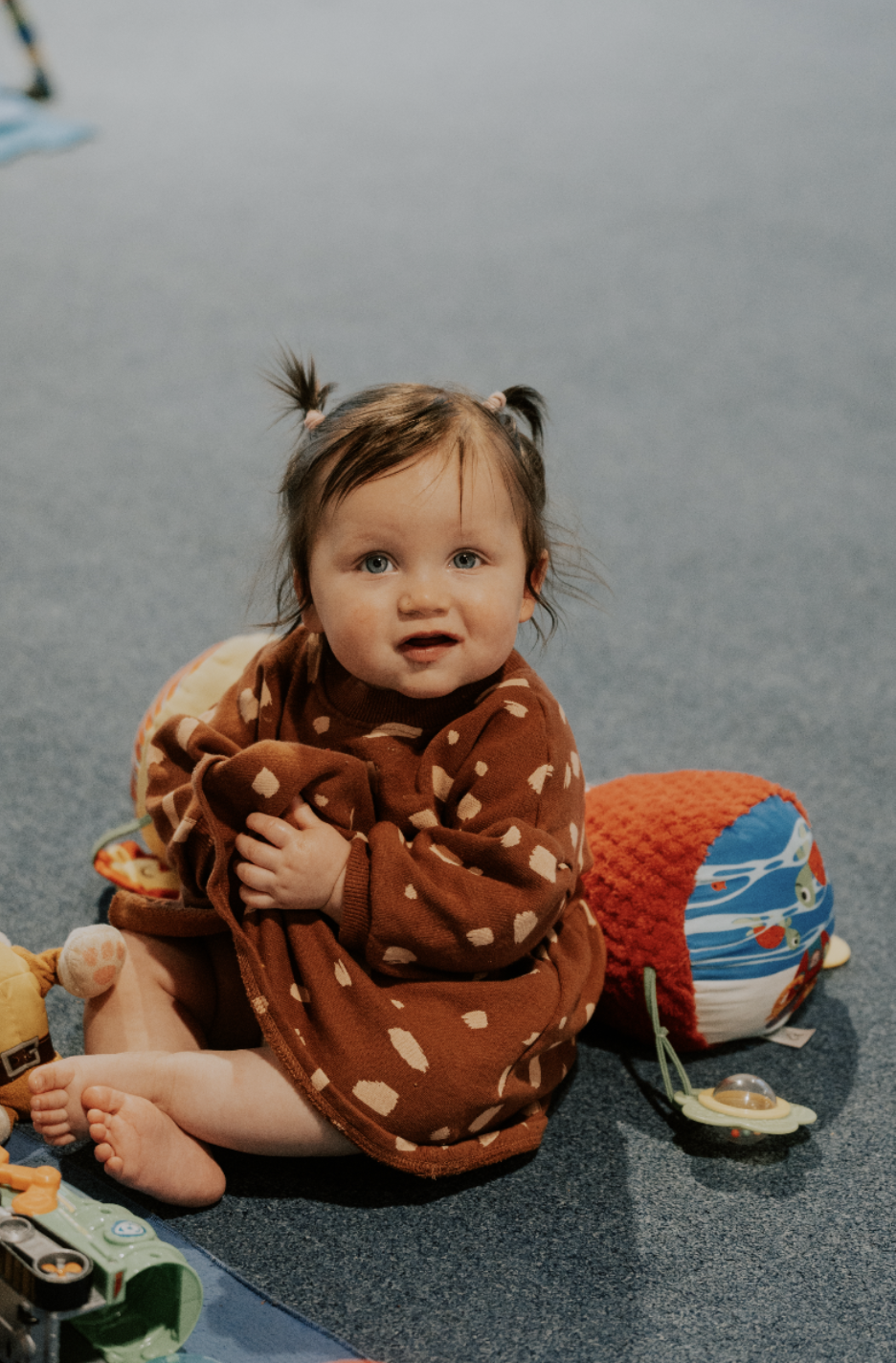 A young child with brown hair in pigtails, wearing a brown onesie with white hearts, sitting on a blue carpeted floor surrounded by toys, looking at the camera.
