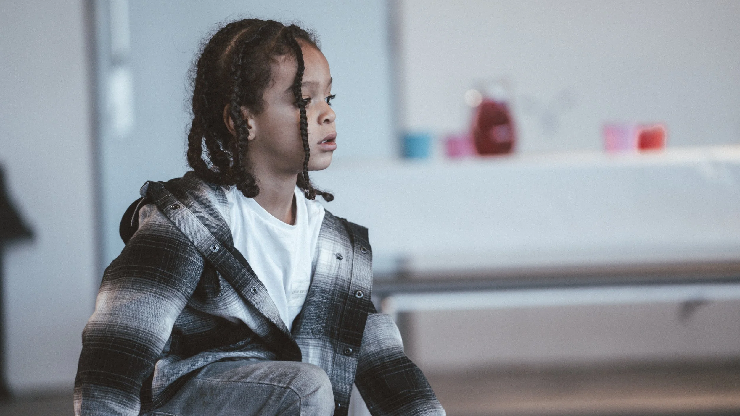 A young girl with curly hair in braids sitting on a chair, wearing a plaid jacket over a white shirt, in a modern interior space with a white background and blurred colorful objects in the background.