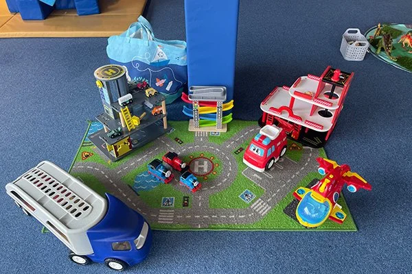 Children's toy playset with a blue and white bus, fire truck, helicopter, city buildings, road, and traffic signs on a green mat.