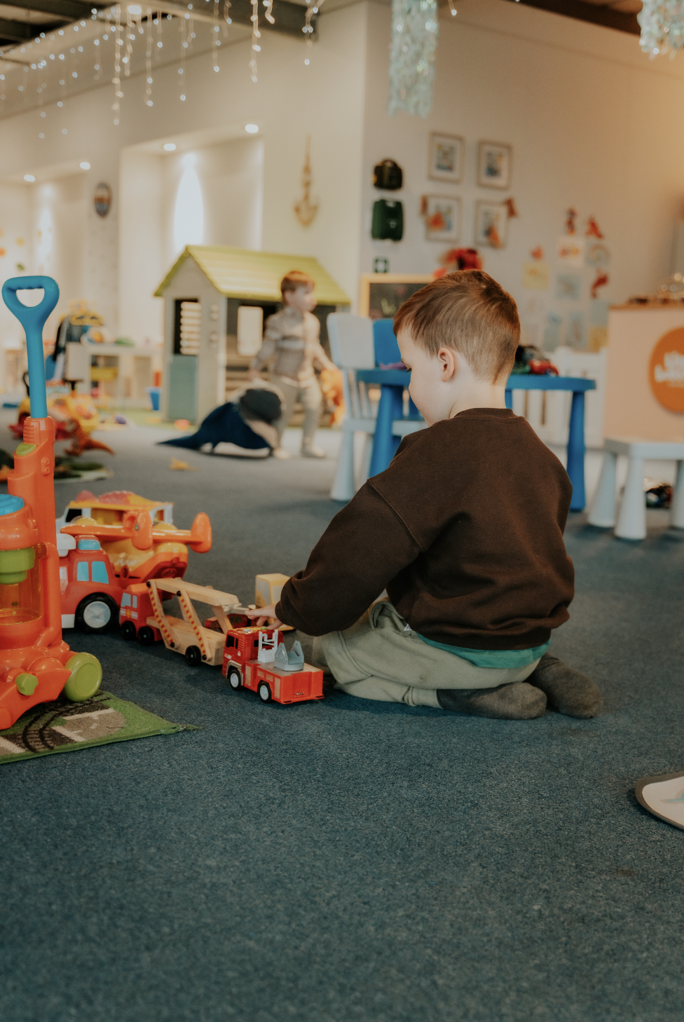 Child playing with toy vehicles, including fire trucks and construction vehicles, on a blue carpeted floor in a children's playroom decorated with fairy lights and artwork.