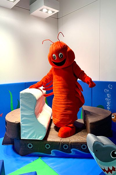 Costumed character of a red lobster with a big smile in an indoor play area with ocean-themed decorations.