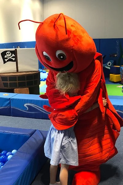 A person dressed in a large, red, crab costume hugging a young girl in a light blue dress. The scene appears to be in an indoor play area with blue padded surroundings and nautical-themed decorations.