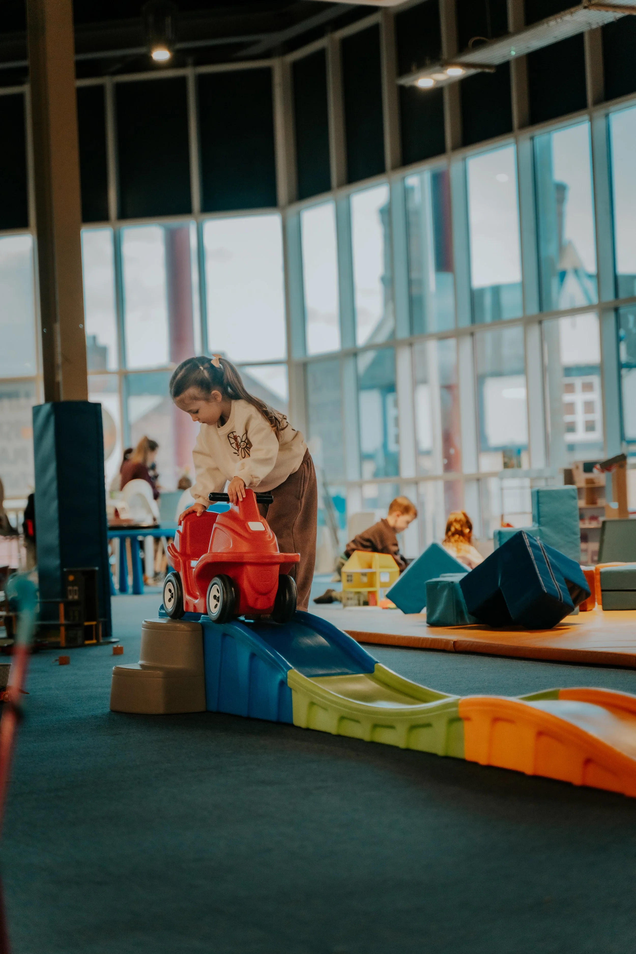A young girl playing with a toy dump truck on a colorful indoor playset, with other children in the background in a bright, spacious play area.