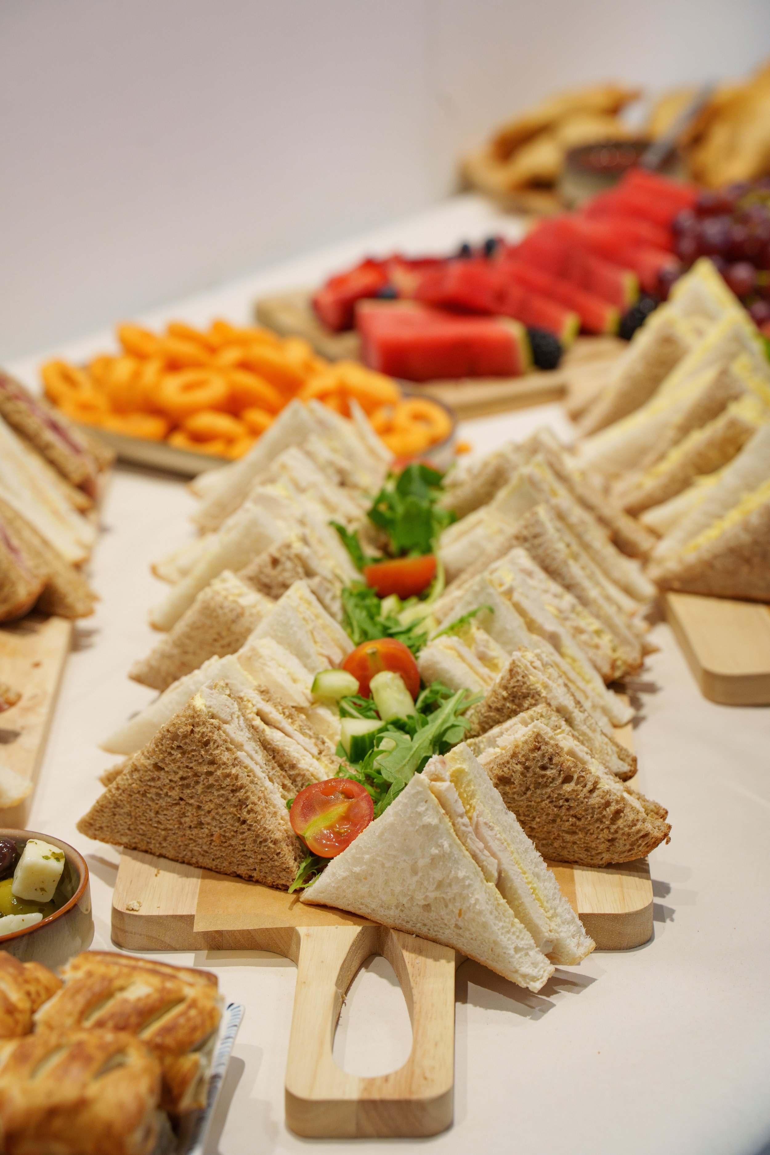 An assortment of finger sandwiches with various fillings arranged on a wooden serving board, garnished with cherry tomatoes and greens, with a background of sliced watermelon and other fruits.