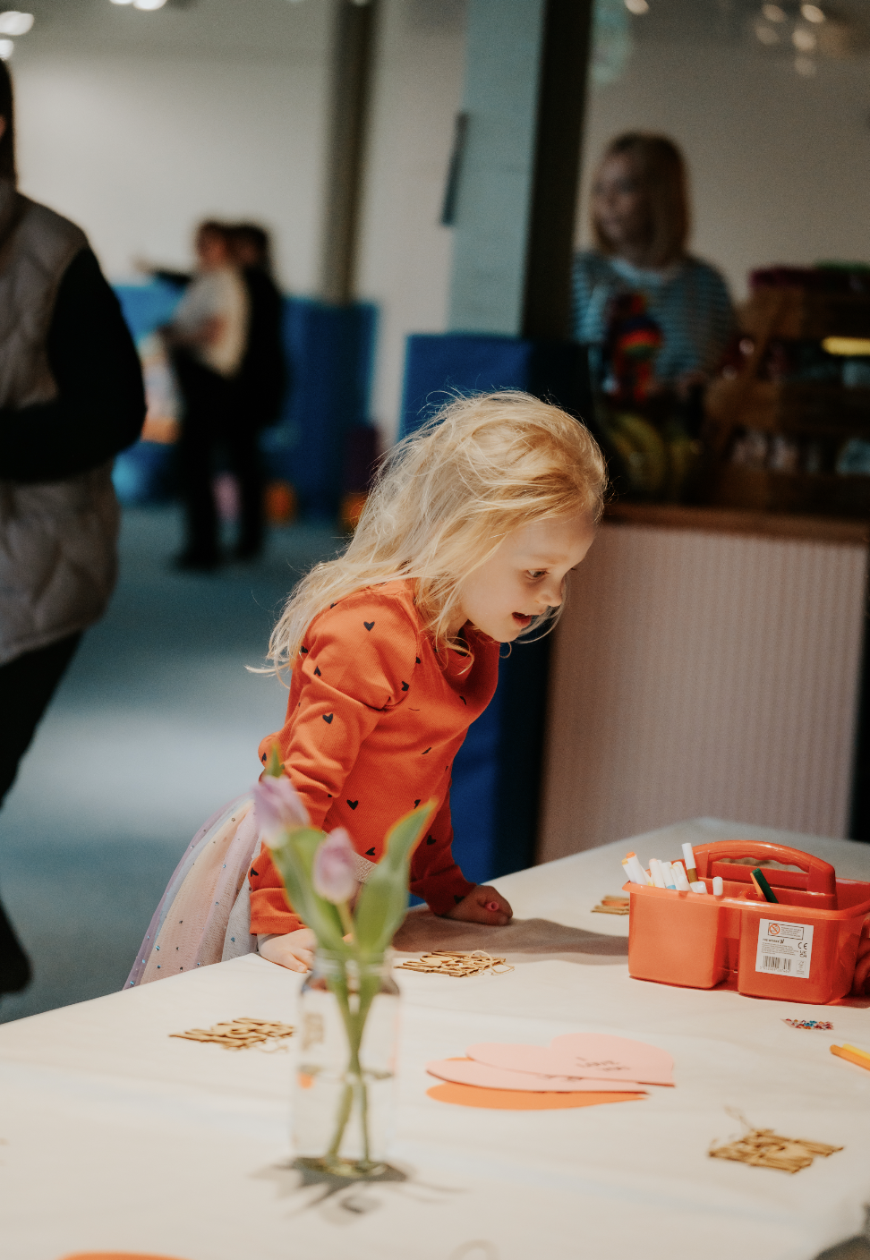 A young girl with blonde hair leaning over a table at a party or gathering, surrounded by paper hearts and art supplies on the table.