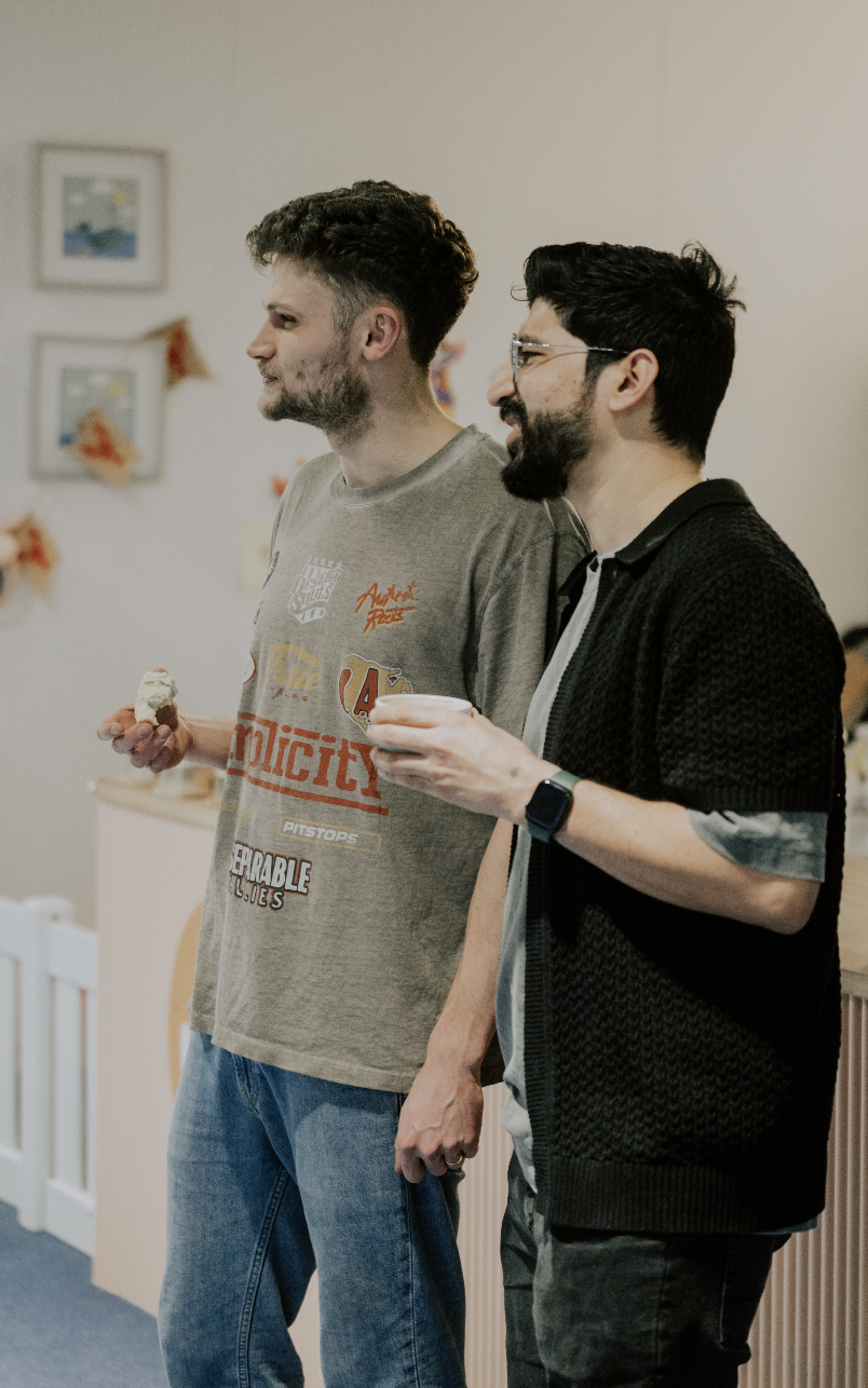 Two men standing indoors, one holding a piece of cake and the other a cup, engaged in conversation.