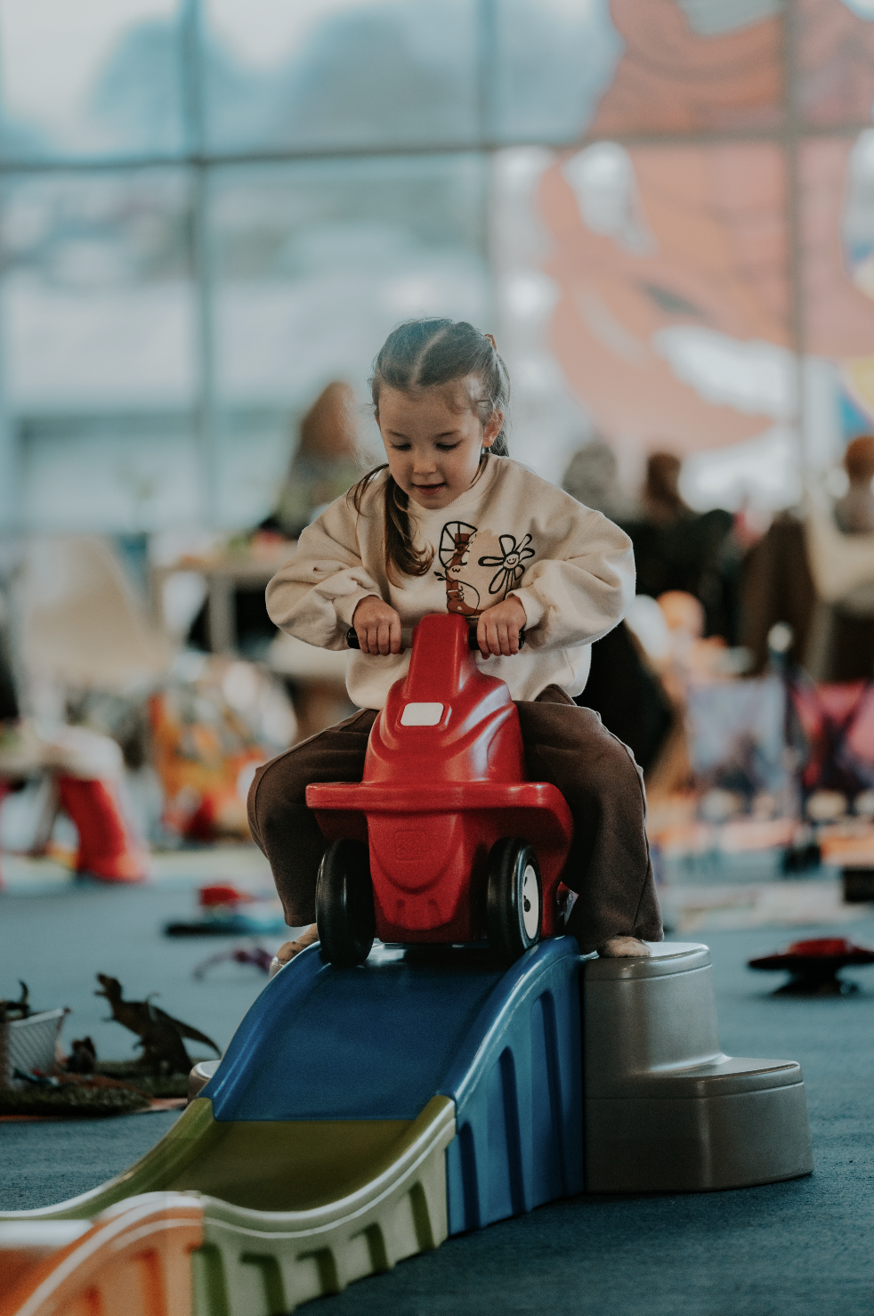 Young girl playing on a red ride-on toy car on a small slide at an indoor play area.