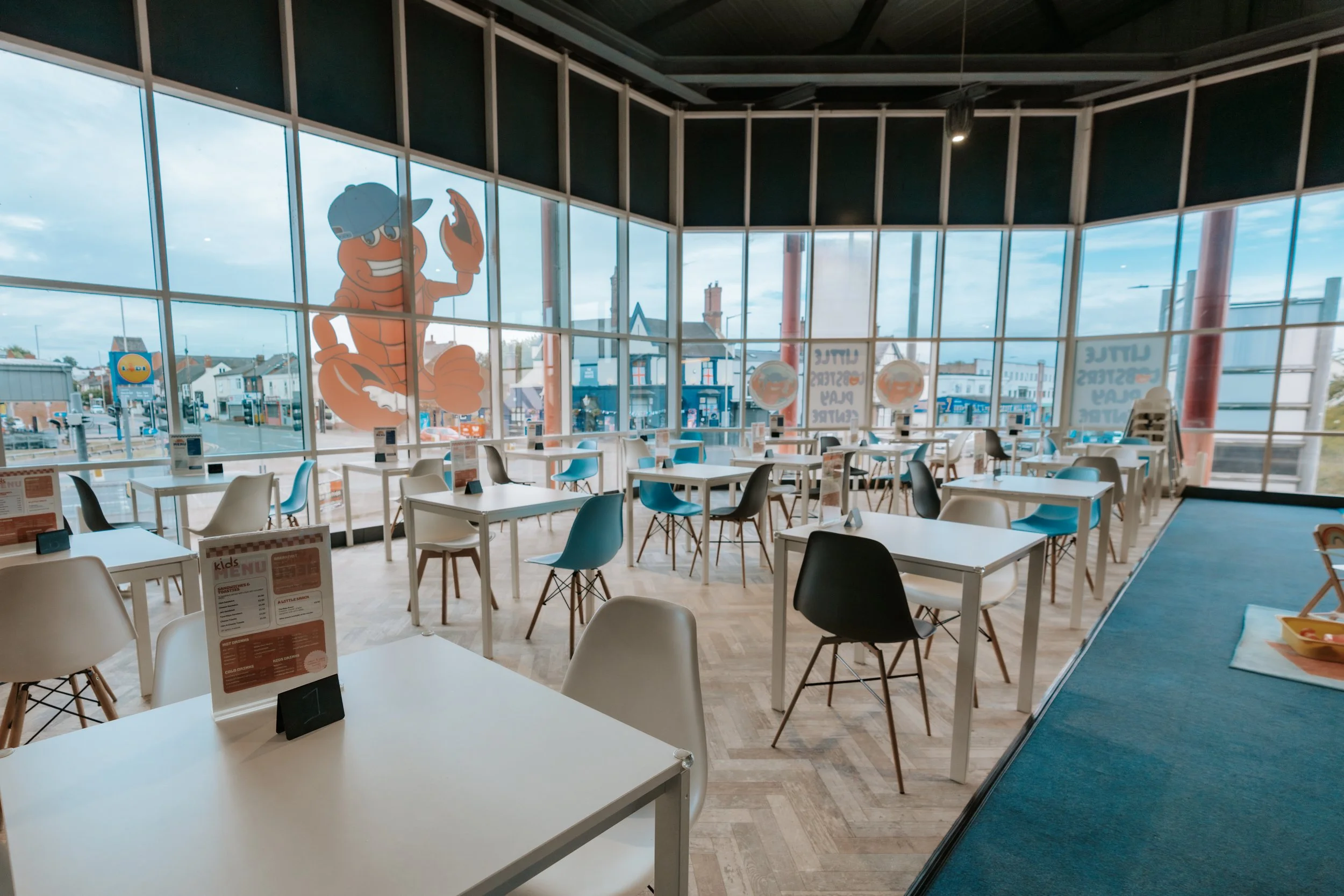 Inside a modern, empty fast-food restaurant with large windows and a view of the street. The windows feature a cartoon lobster wearing a construction helmet. The interior has white tables, blue and white chairs, and light wood flooring.
