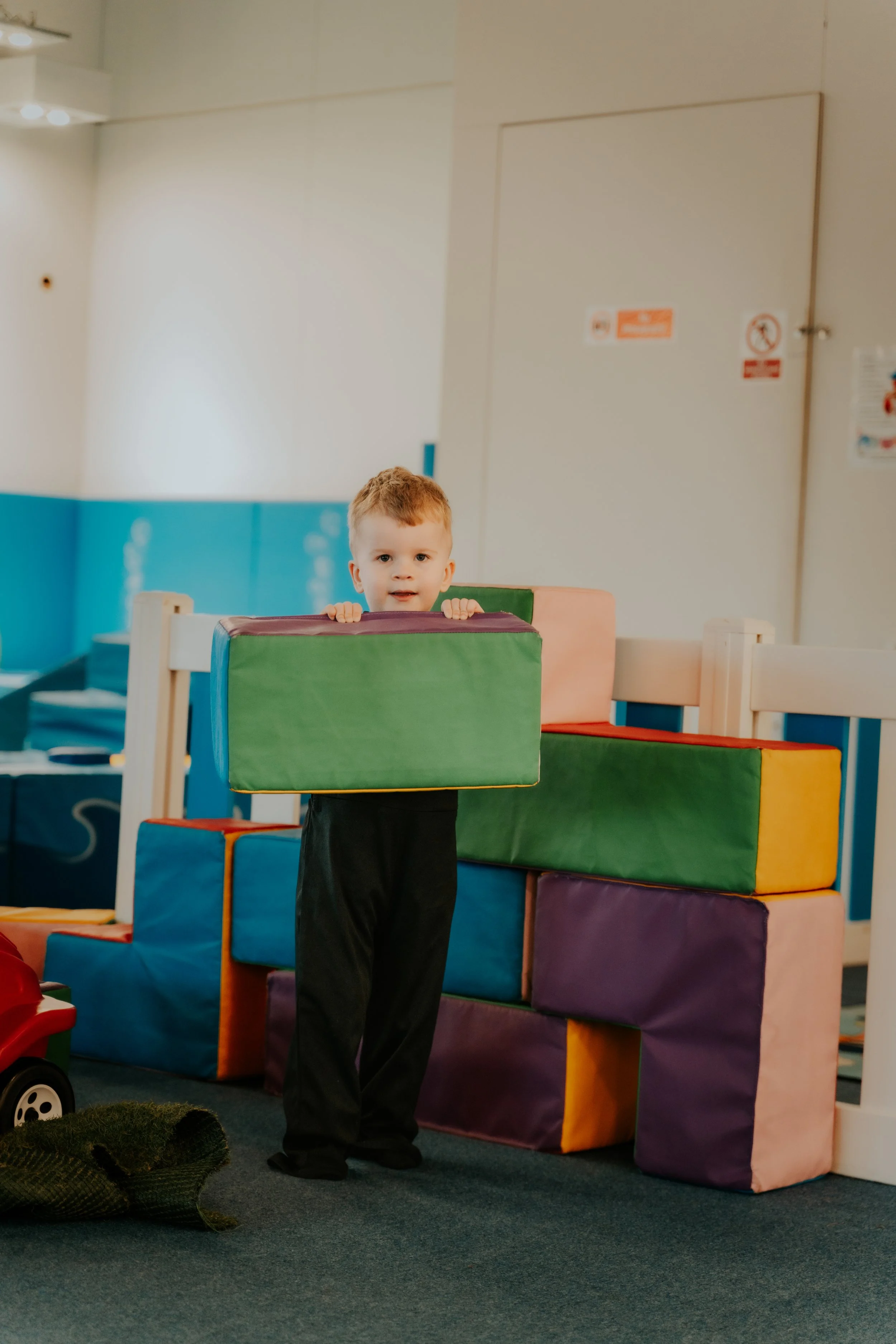 A young boy with red hair holding a large foam block in a playroom with colorful foam blocks stacked behind him.
