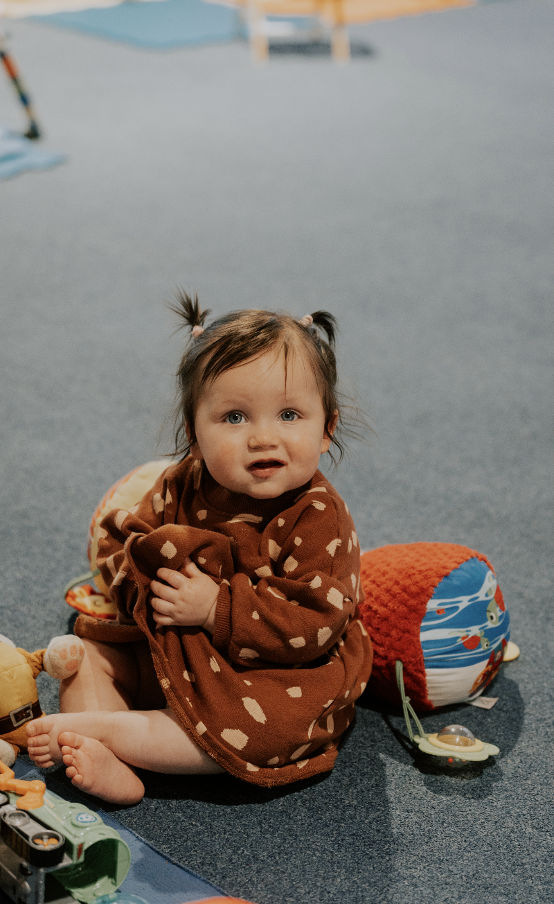 A young child with brown hair tied in pigtails, sitting on a blue carpeted floor, wearing a brown sweater with white polka dots, surrounded by toys including a plush ladybug, a pacifier, and other colorful toys.