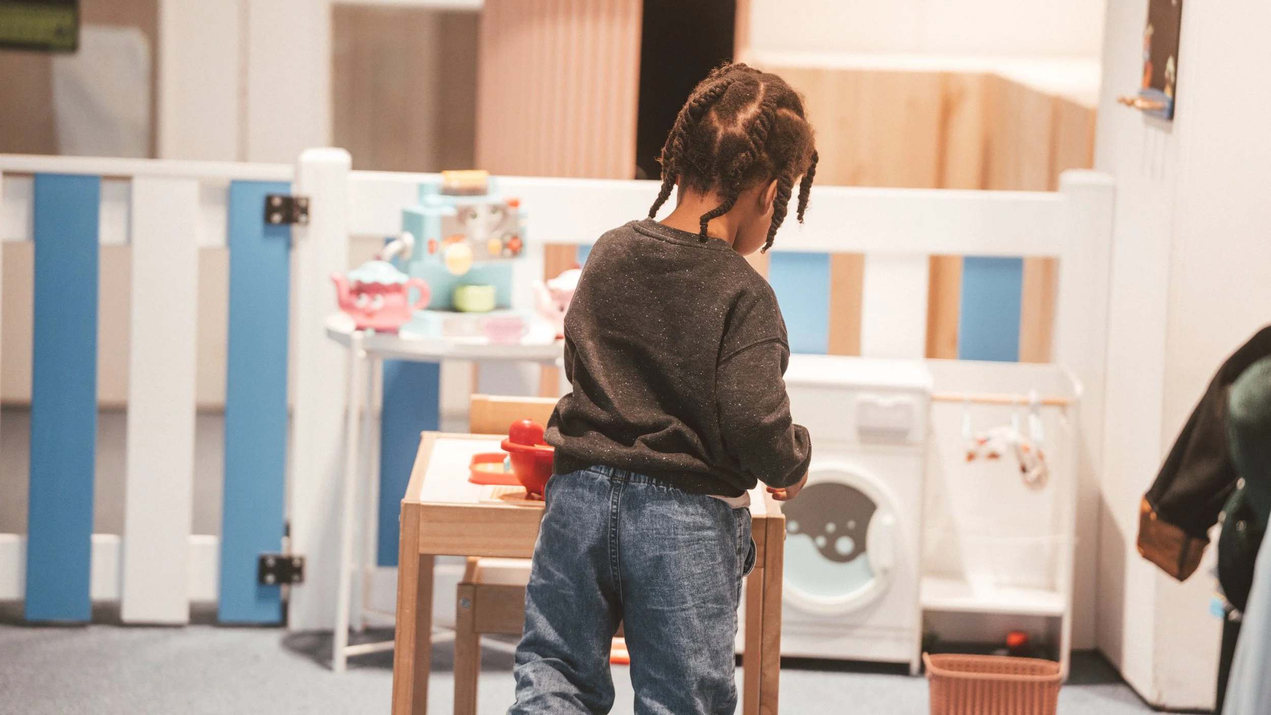 A young girl with braided hair wearing a dark long-sleeve shirt and jeans, standing in a room with a toy kitchen set, a washing machine, and a laundry basket.