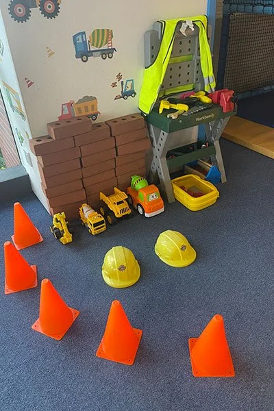 Construction toy area with orange traffic cones, yellow toy vehicles, hard hats, toy bricks, and a high-visibility vest on a shelving unit.