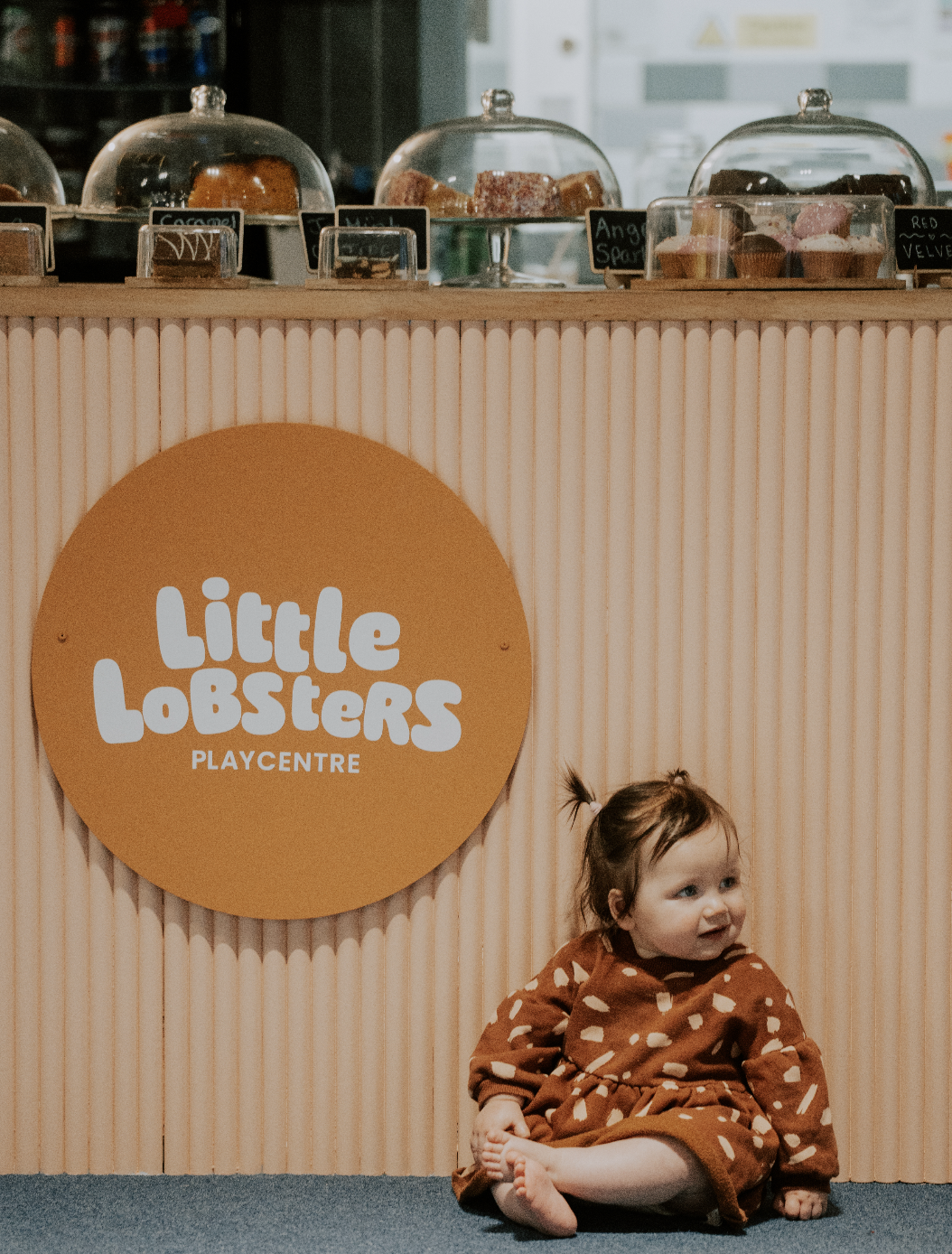 A young girl sitting on the floor at the Little Losters play center, with a wooden counter behind her displaying baked goods under glass covers and a large circular sign with the play center's name.
