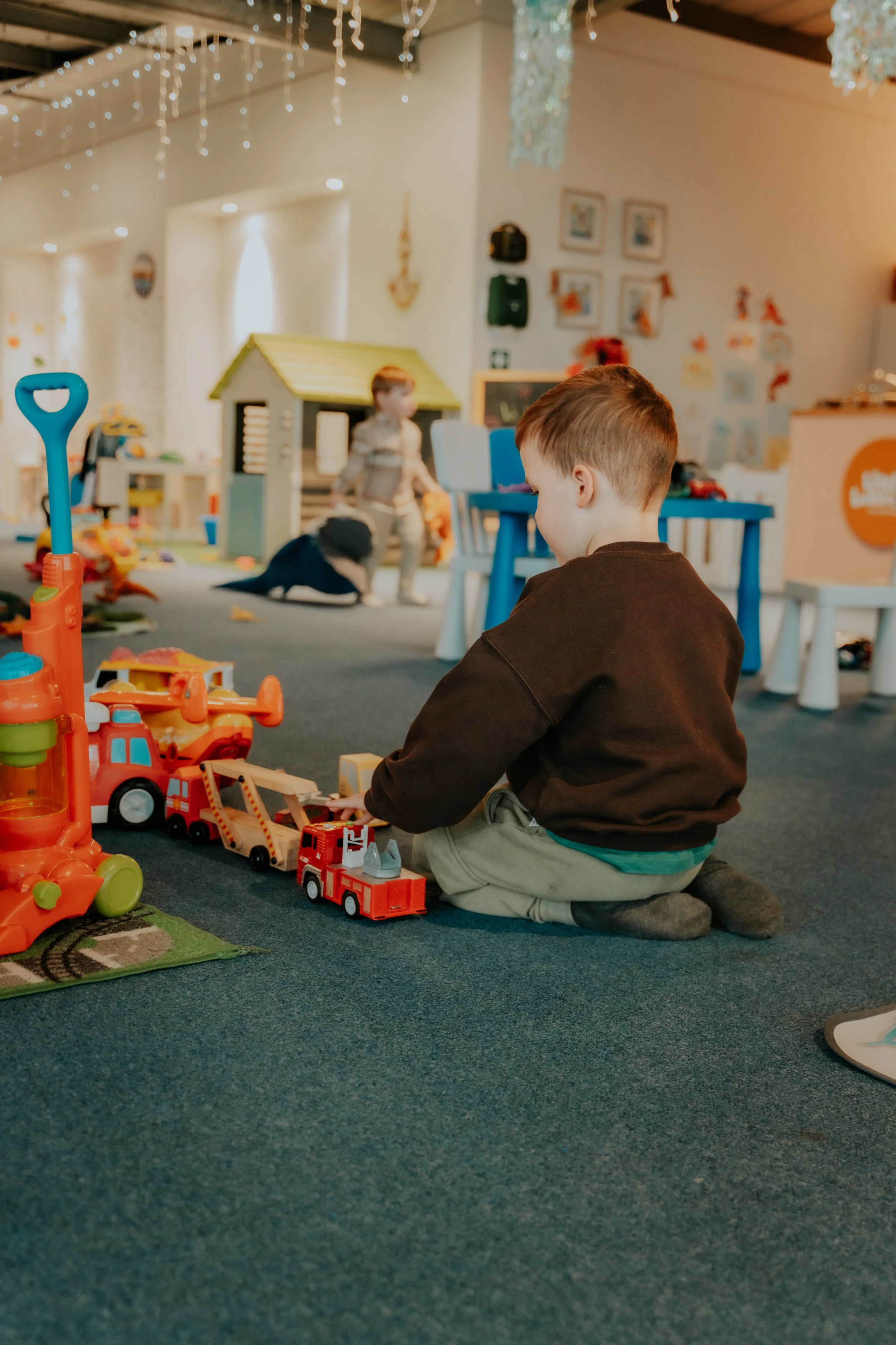 Child sitting on a carpeted floor playing with toy fire trucks and other toys in a playroom decorated with string lights, artwork, and a playhouse in the background.