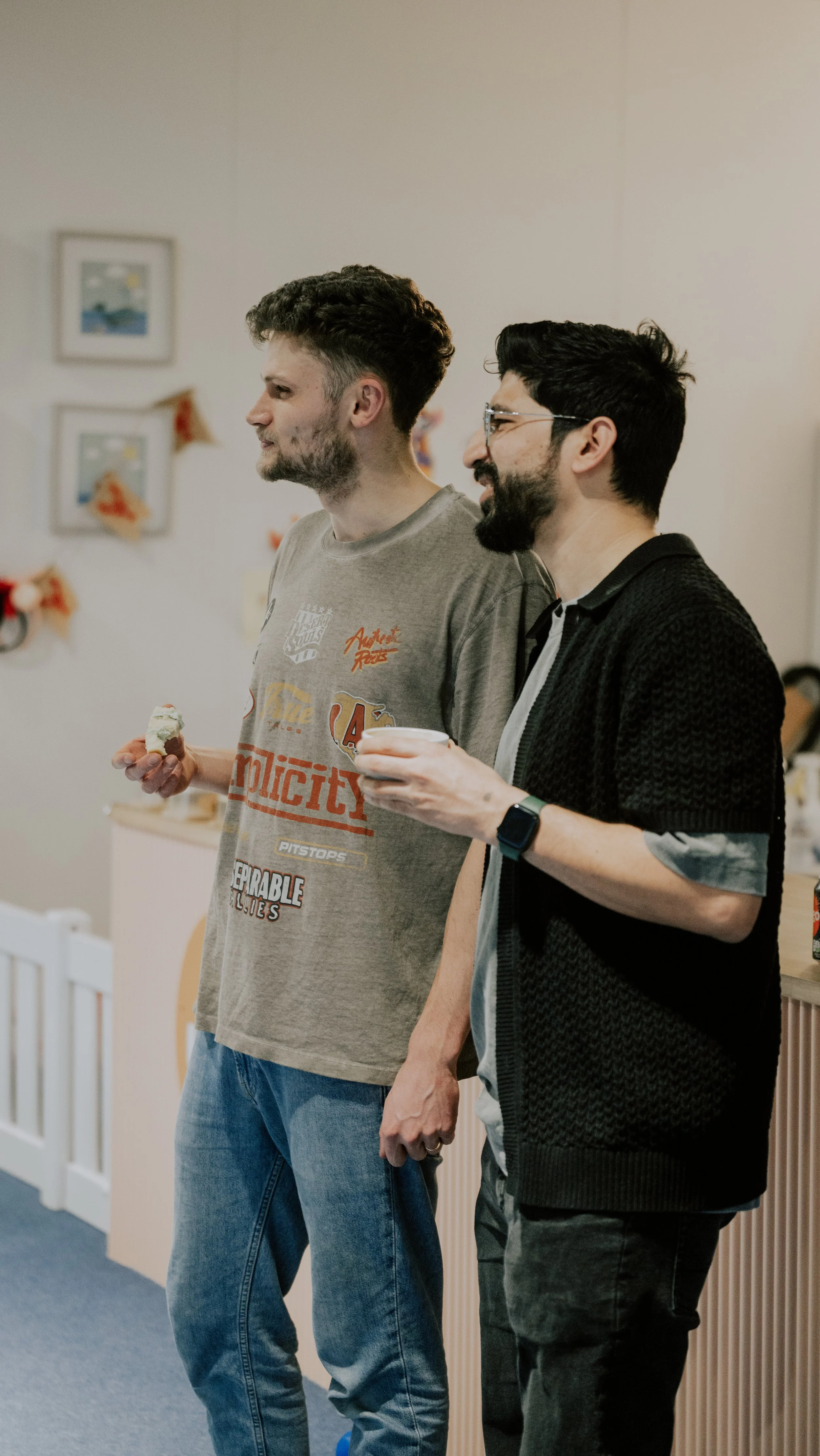 Two men standing indoors, one holding a cup and the other holding a small piece of food, possibly a cupcake.