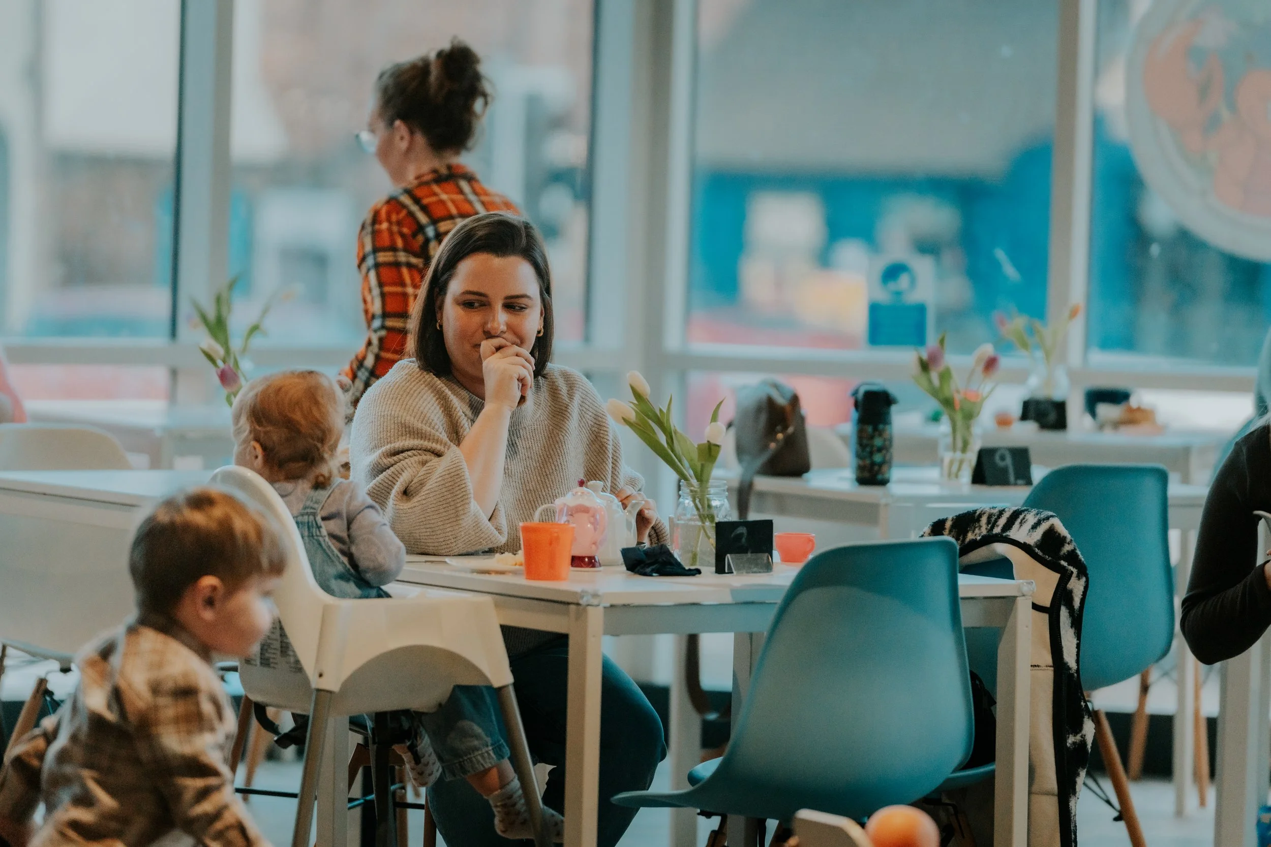 A woman with dark hair sitting at a table with children, one in a striped sweater, with tulips in a vase on the table, in a bright room with large windows.