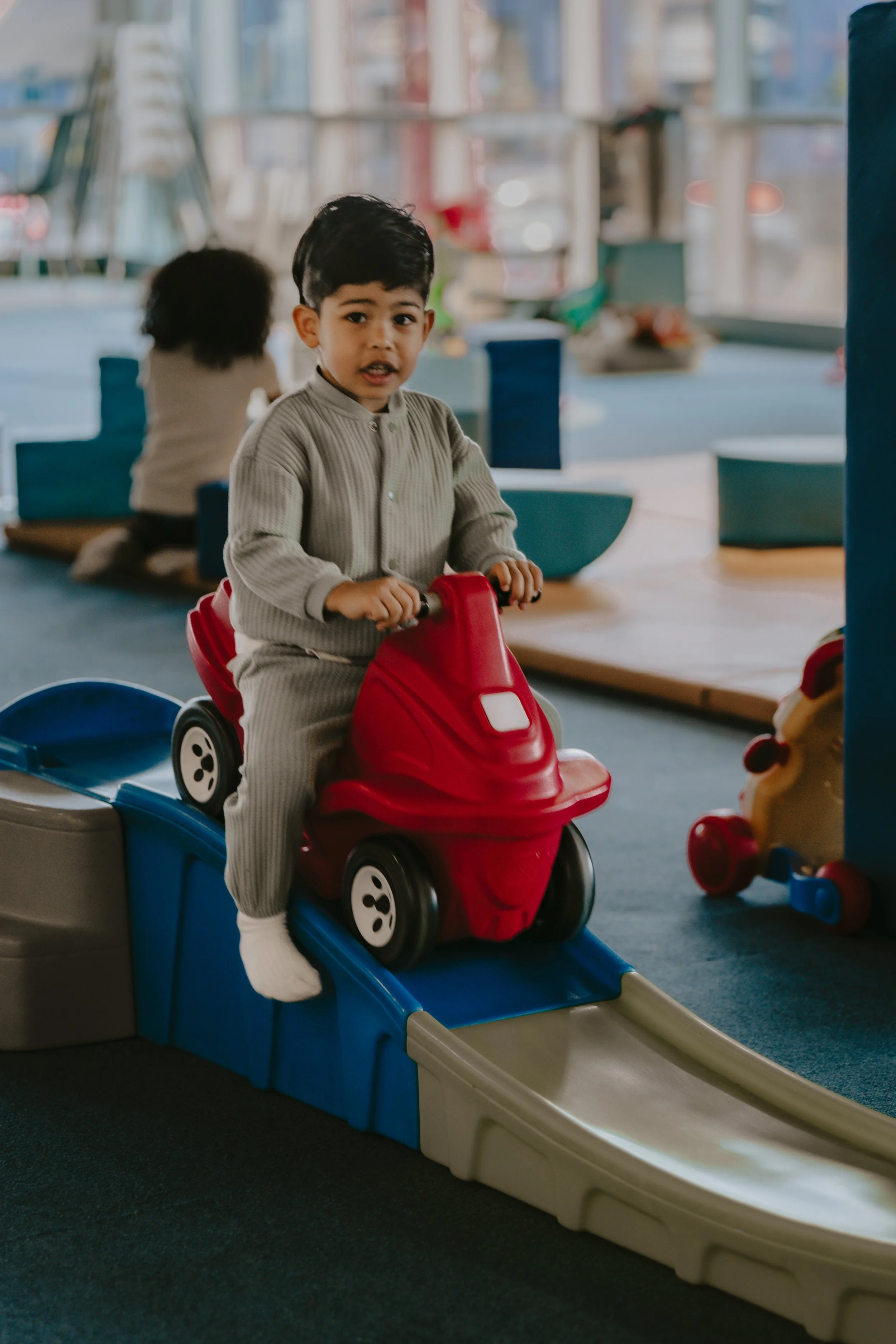 A young boy with dark hair and a gray sweater riding a red and blue toy car on an indoor slide, with a blurred background of a play area and large windows.