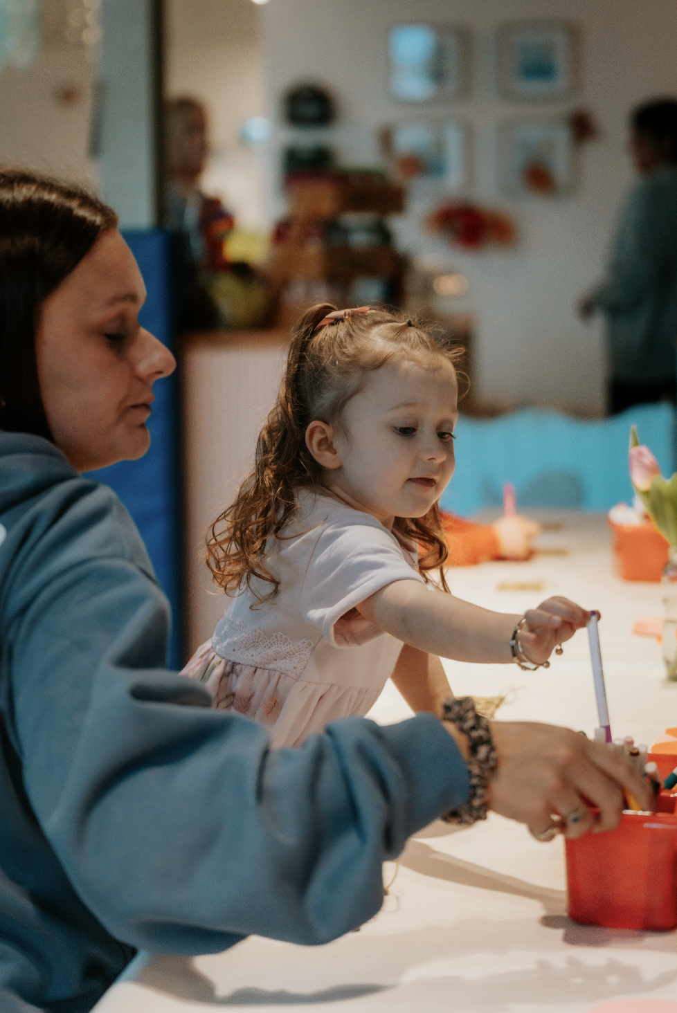 A young girl with curly hair and a pink dress is playing with art supplies at a table, assisted by a woman. In the background, other people are present in a cozy indoor setting decorated with pictures and colorful items.
