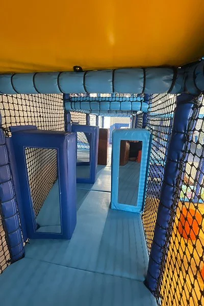 Interior view of an indoor playground with padded blue flooring, netting on the sides, and blue foam blocks with mirrors.