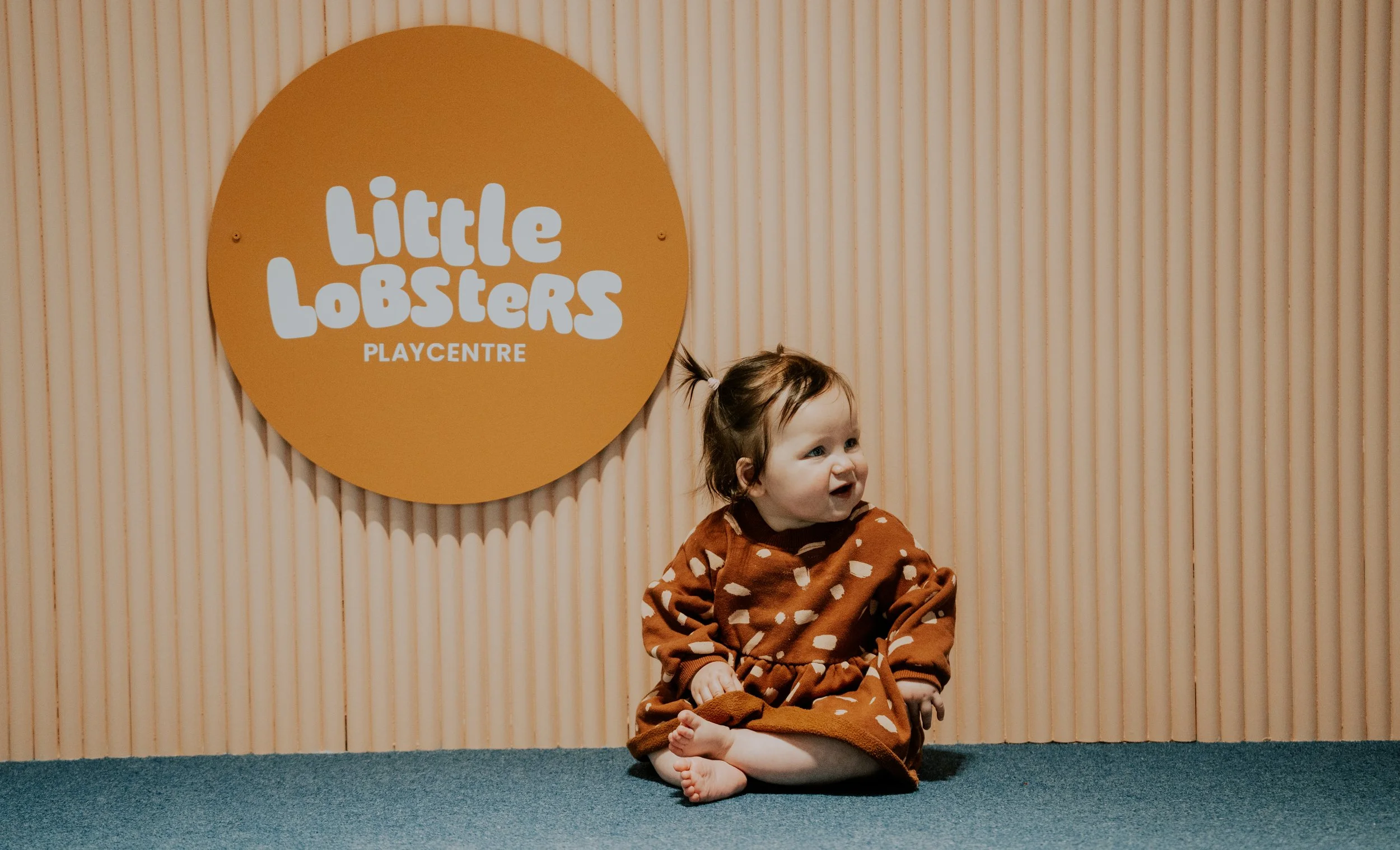 A young child with brown hair tied in a small ponytail, wearing a brown dress with white polka dots, sitting on a blue carpeted floor in front of a vertical wooden panel wall and a large round sign that reads 'Little Lobsters Playcentre' in white tex