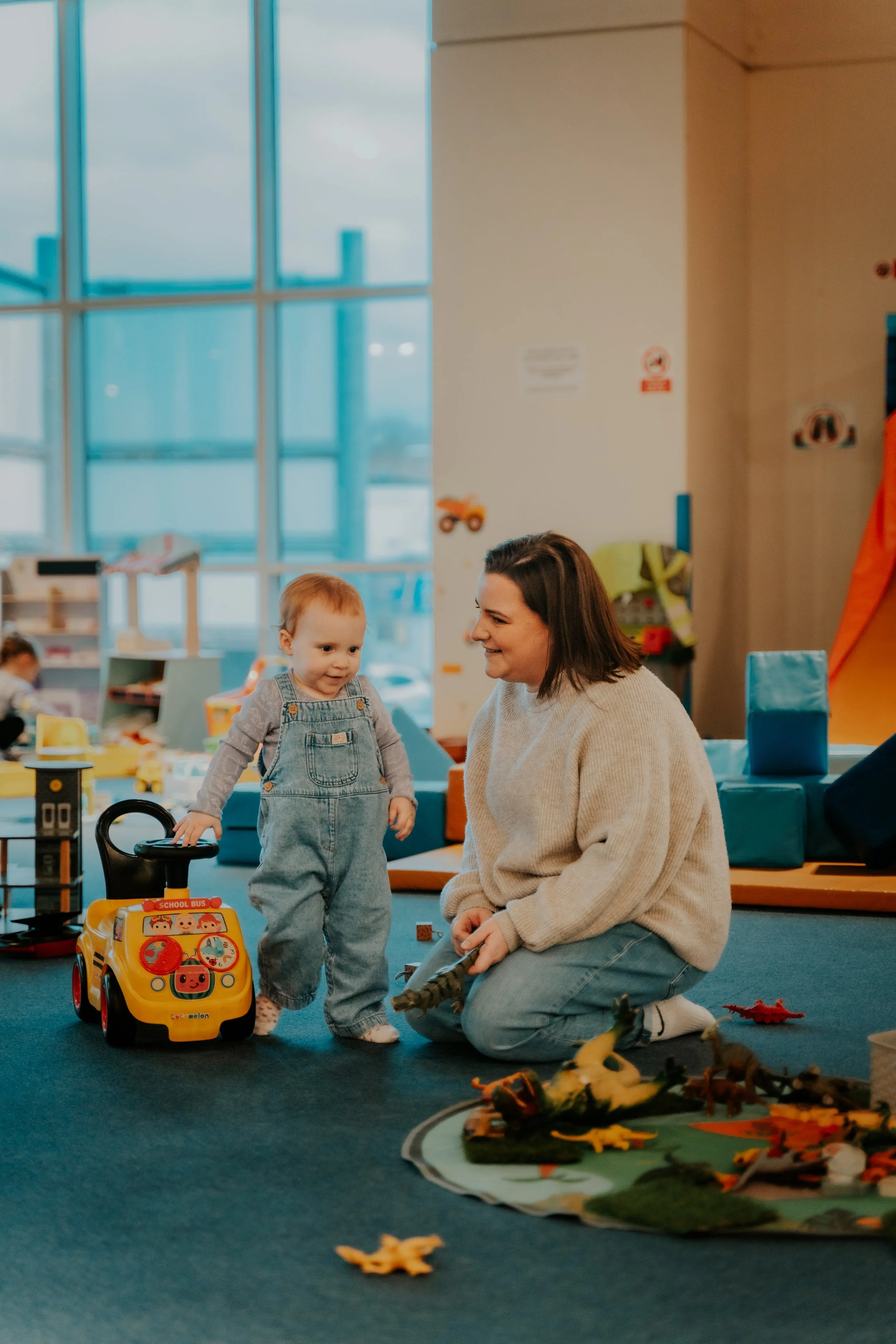A woman and a young boy playing with toys in a playroom with large windows and colorful decorations.