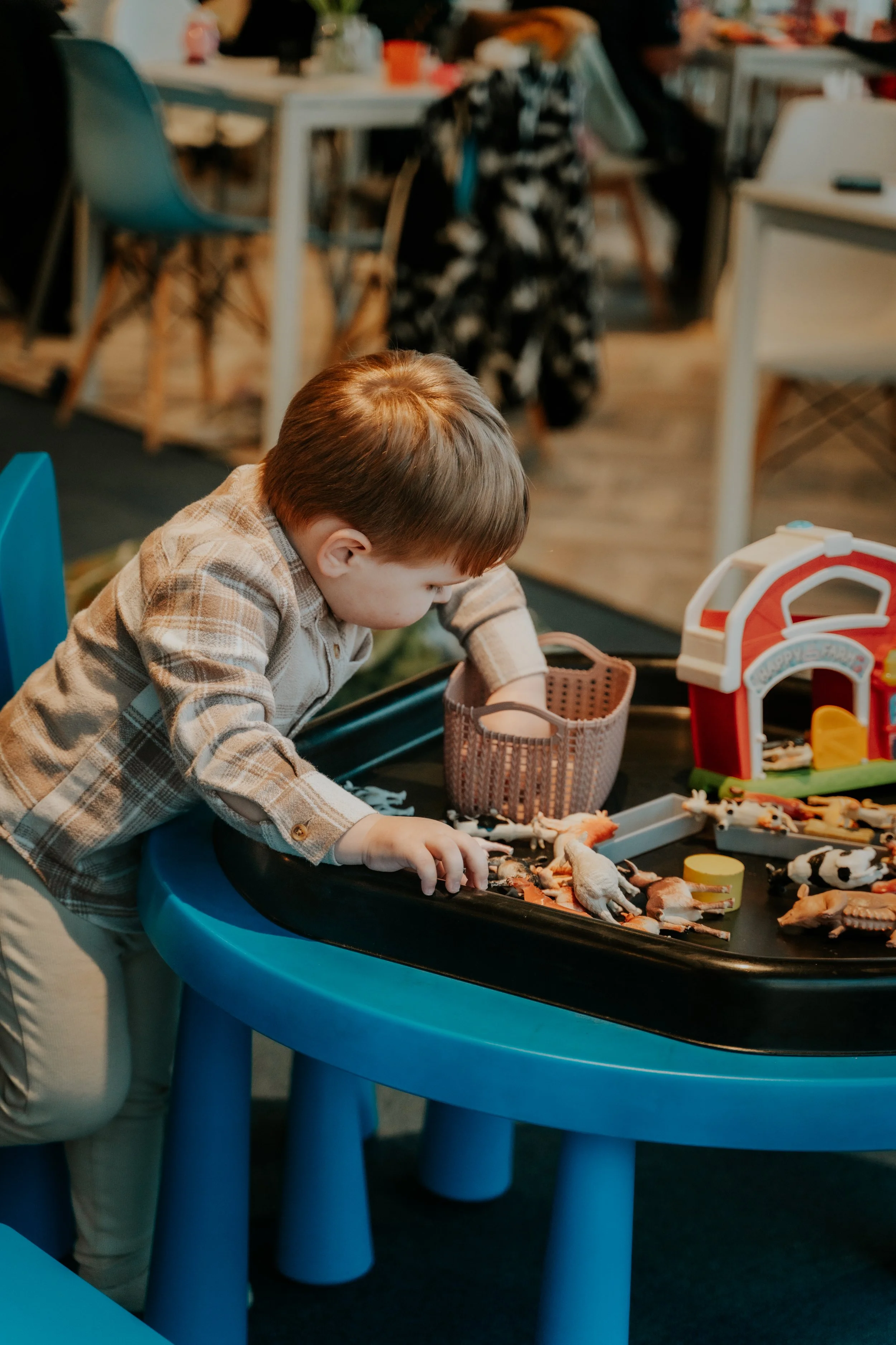 A young boy with brown hair wears a plaid shirt and beige pants, leaning over a black and blue play table with toy animals and objects, in a room with chairs and tables, some blurred figures and supplies in the background.