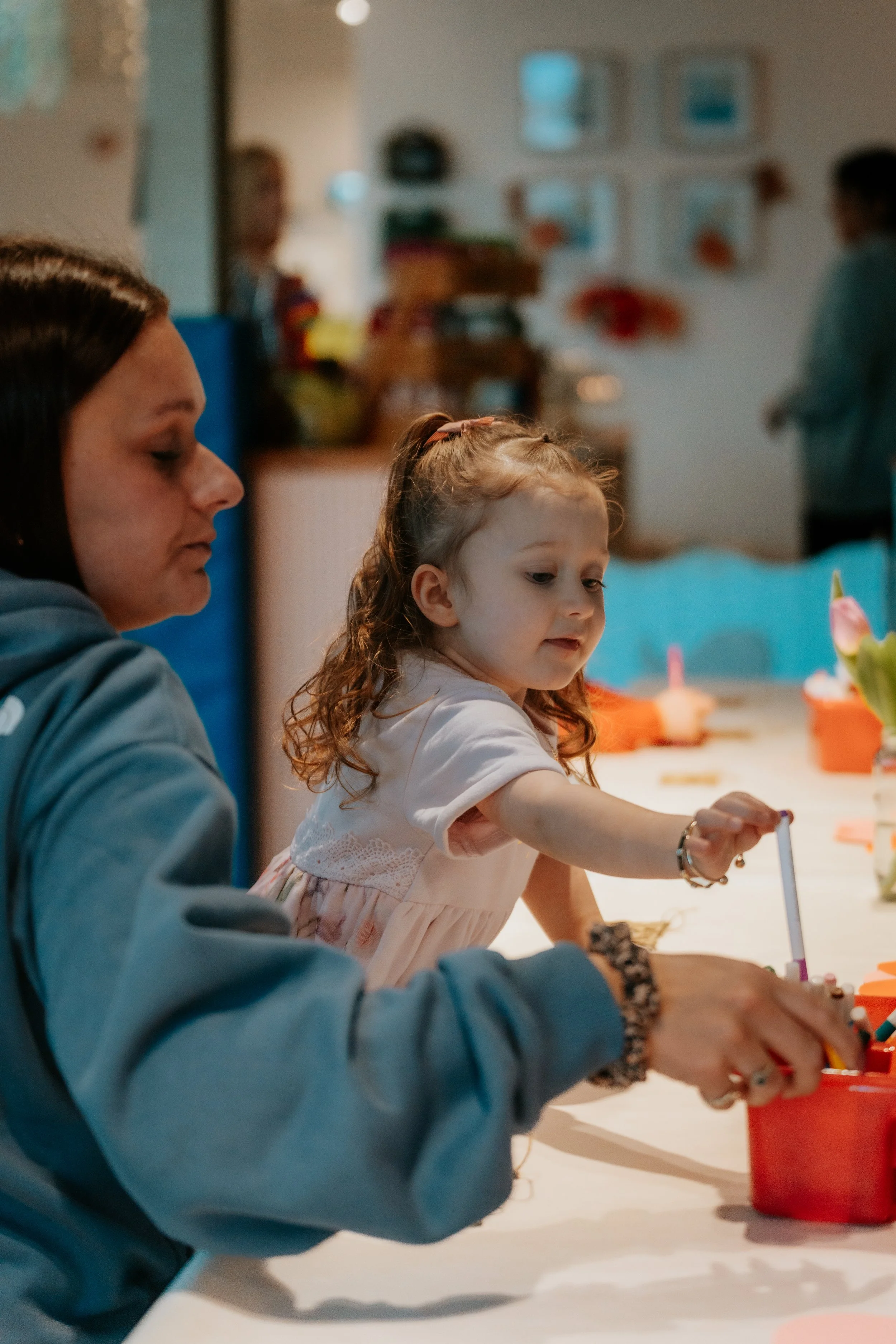 A woman and a young girl sitting at a table, engaged in arts and crafts activities. The girl is reaching into a red container, while the woman looks on. In the background, other people are visible in a cozy, decorated room.