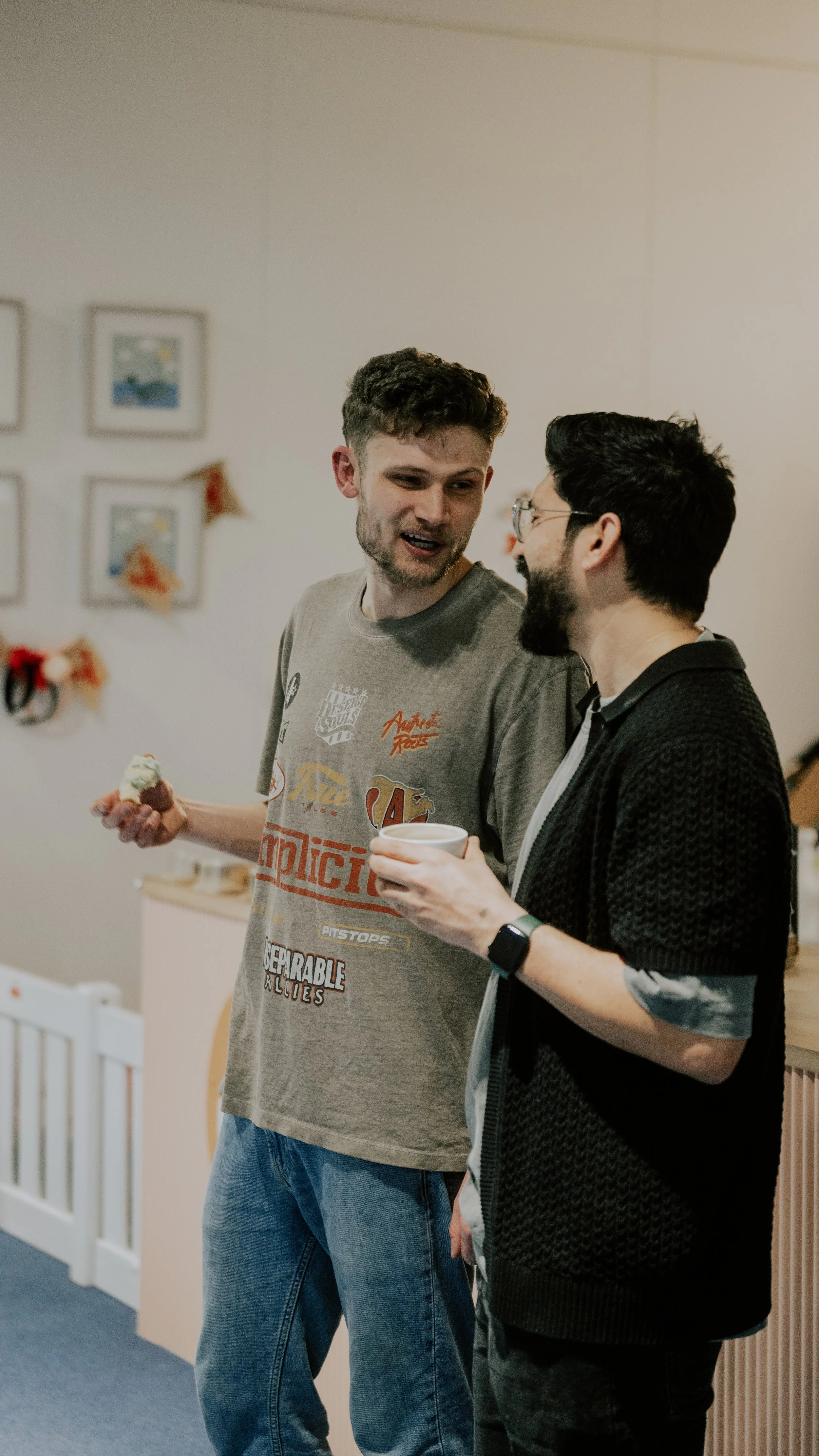 Two men engaged in conversation at a casual gathering, one holding a cupcake and the other holding a cup, in a room decorated with framed pictures and holiday decorations.