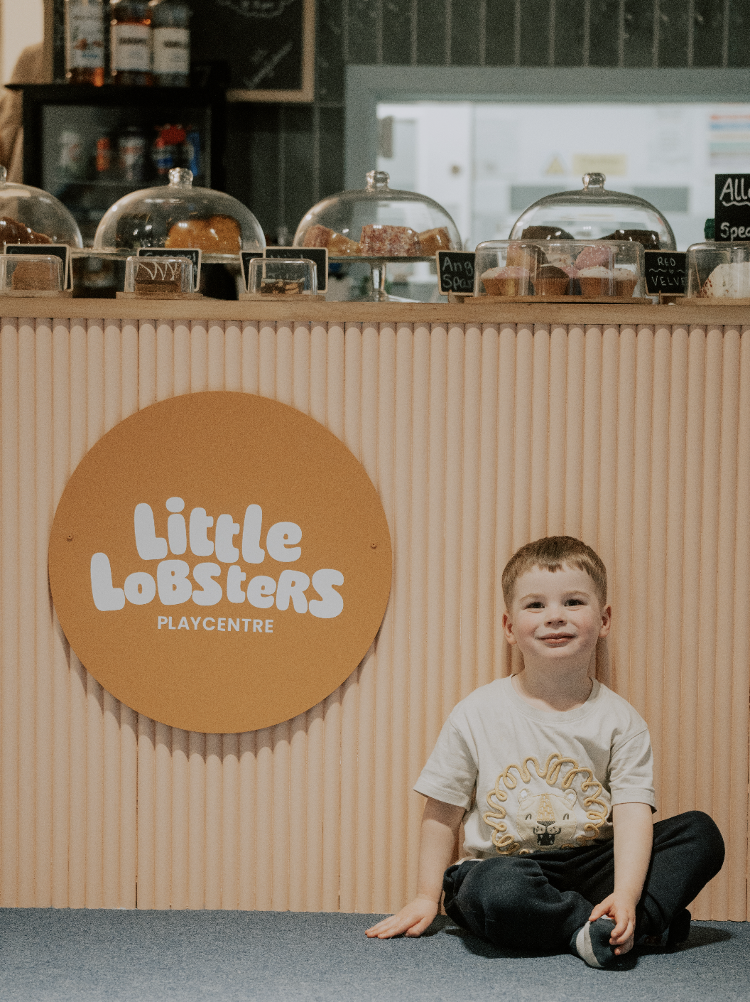 A young boy sitting on the floor in front of a display counter at Little Lobsters Playcentre, with baked goods under glass covers visible behind the counter.