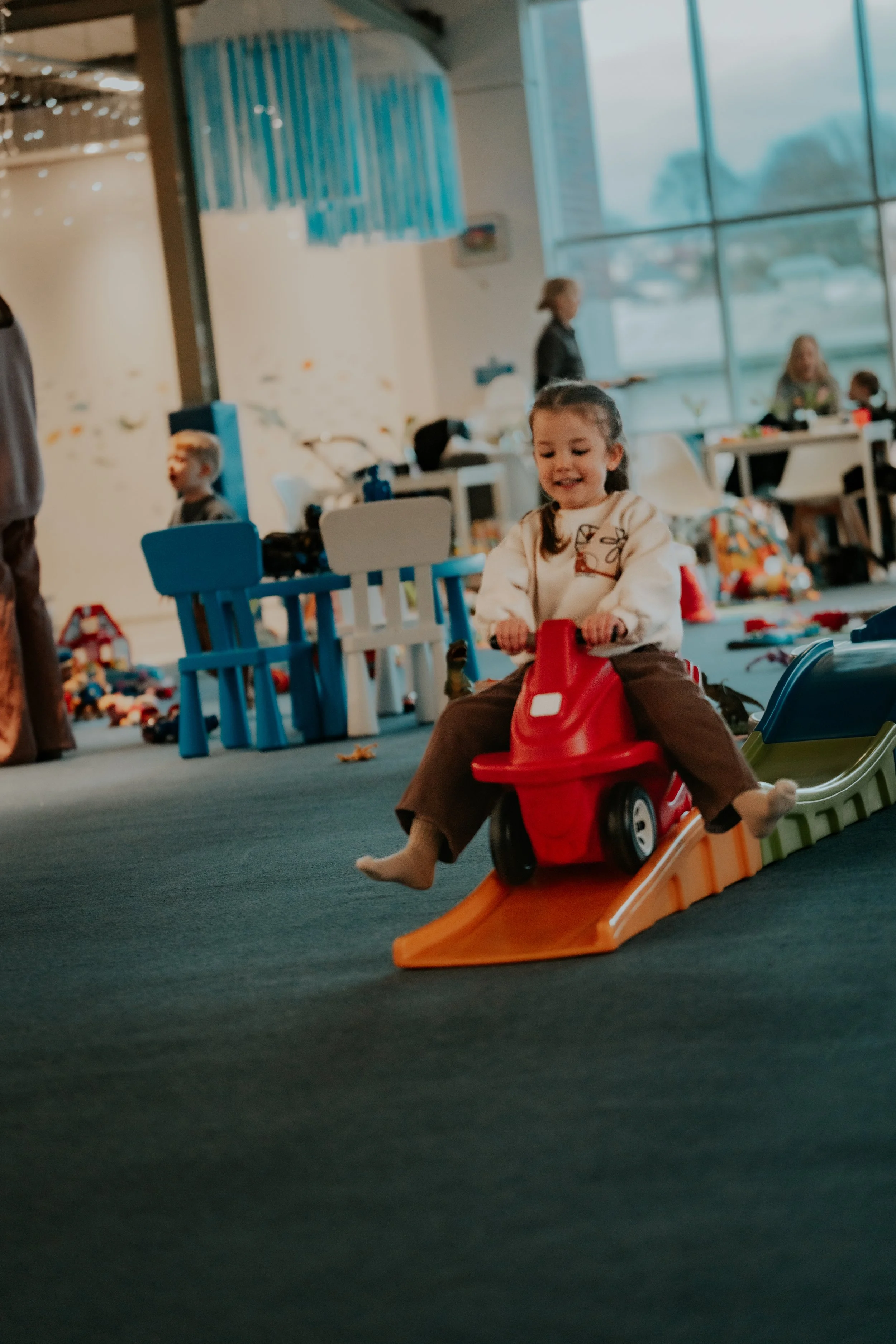 A young girl with braided hair riding a small orange toy car with a slide inside an indoor play area, with toys and children in the background.