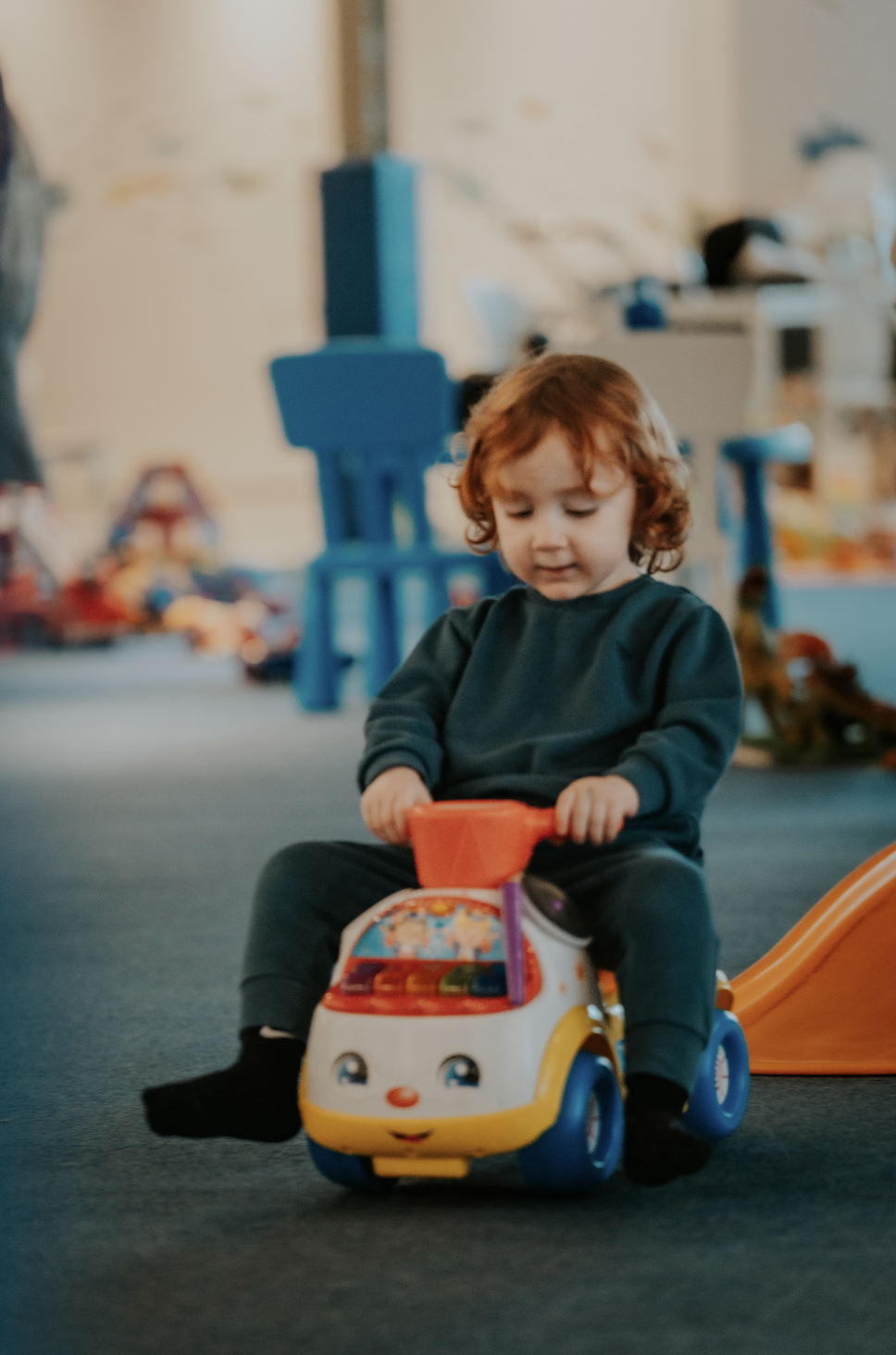 A young child with curly red hair sitting on a colorful ride-on toy vehicle in a playroom.