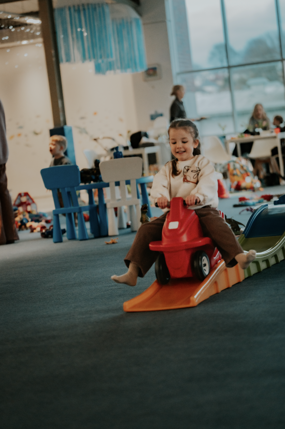 A young girl with dark hair riding a red toy car down a plastic ramp in an indoor playground or play area, with other children and adults in the background.