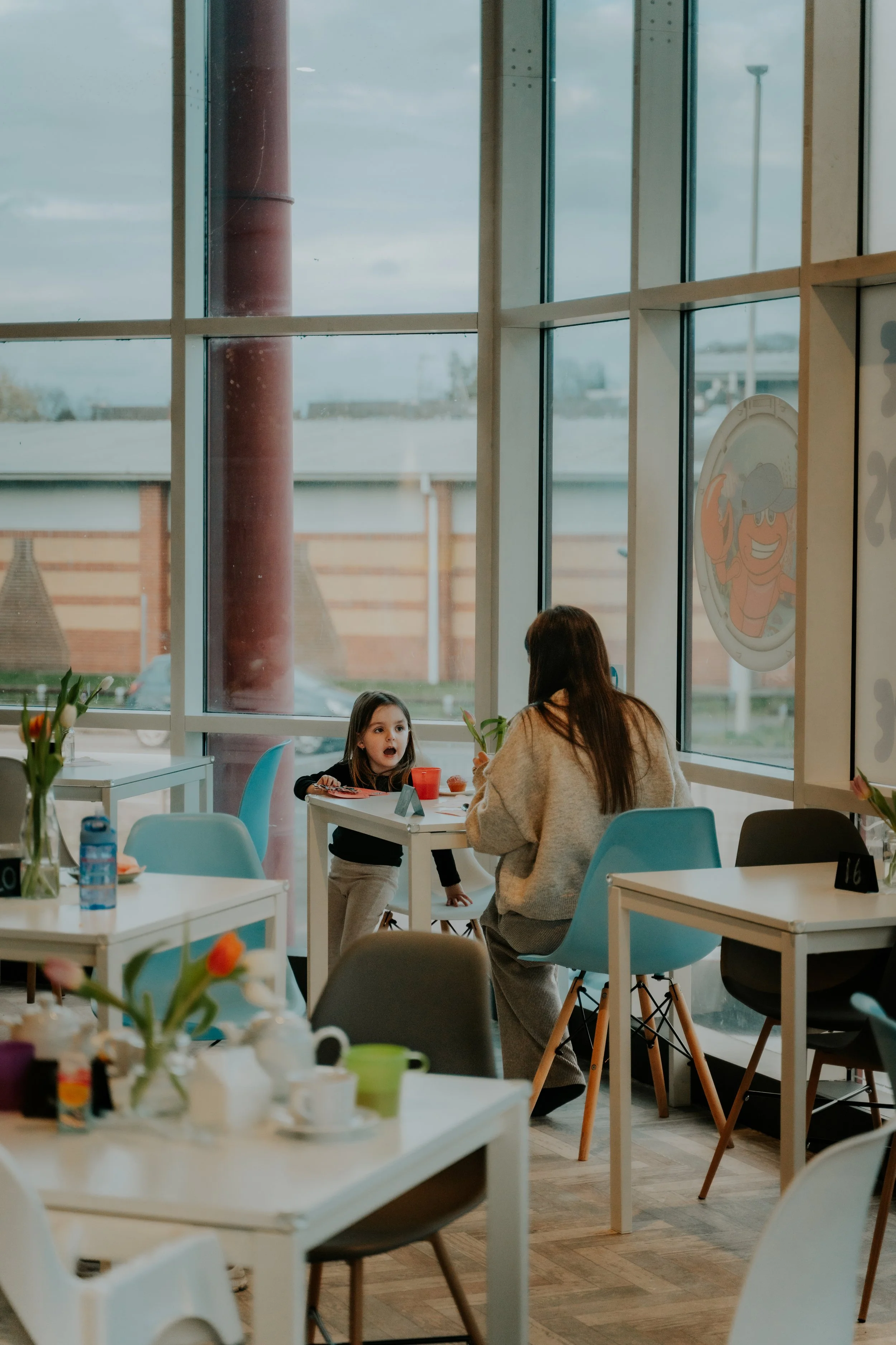 A woman with long brown hair sitting across from a young girl with brown hair at a table in a brightly lit restaurant or cafe. The girl is holding a fork, and there are plates, cups, and a vase with flowers on the table. Large windows reveal a cloudy