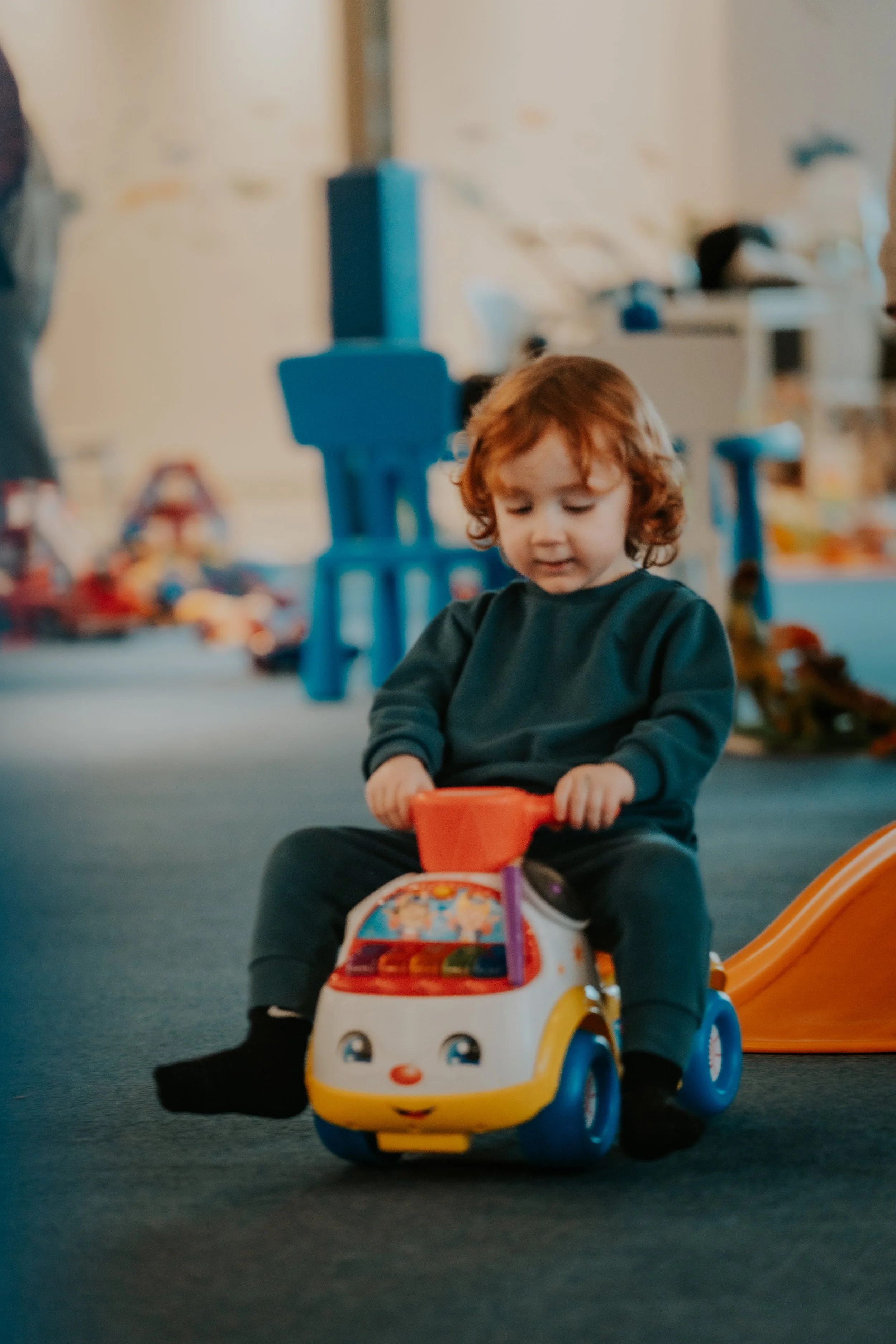 A young boy with curly red hair riding a colorful toy car with a smiling face design in an indoor play area.