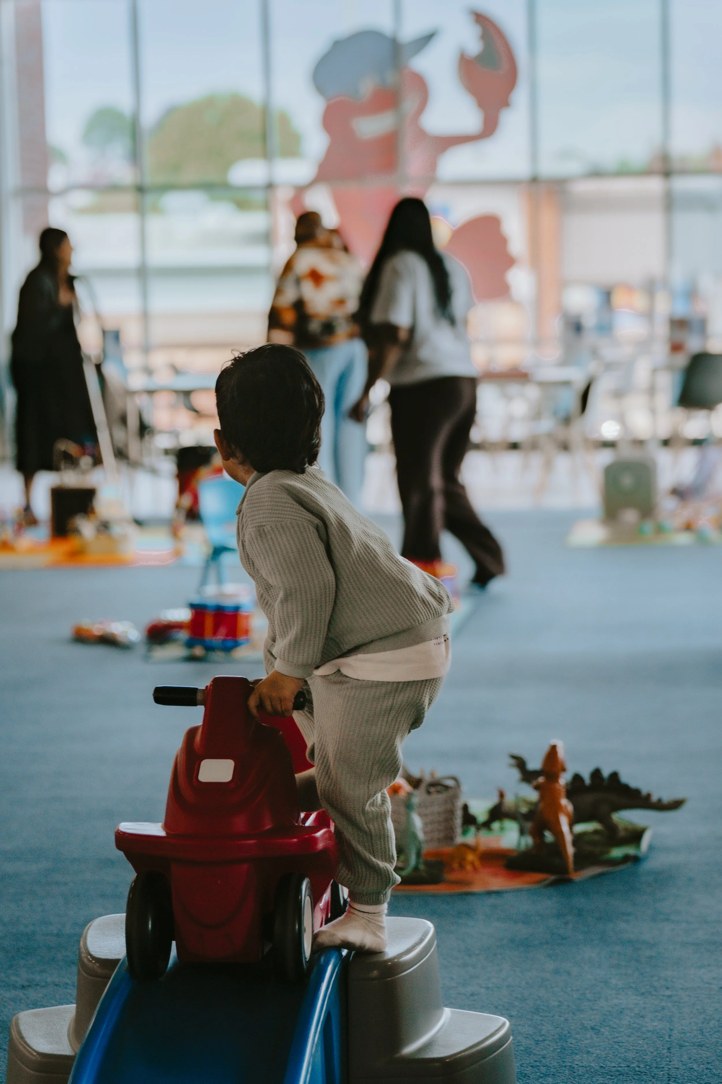 A young child wearing pajamas riding a red toy car on a step in an indoor play area with various toys and figures on the floor, while several adults are talking and socializing in the background near large windows.