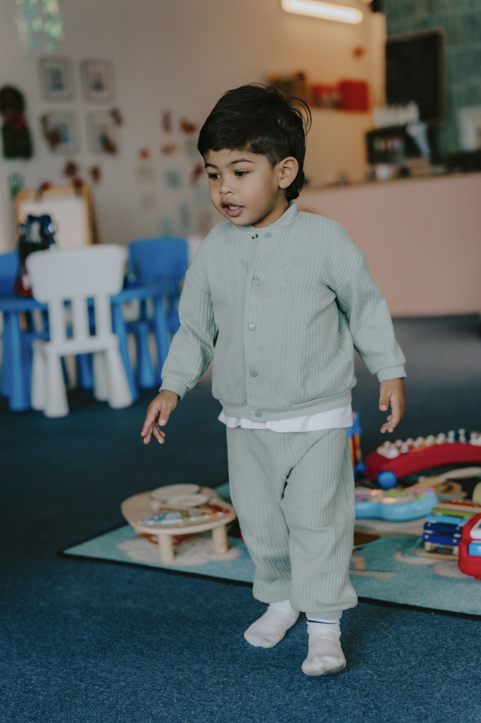 A young boy with dark hair and tan skin walking in a playroom or classroom surrounded by toys and furniture, including blue chairs and a play rug.