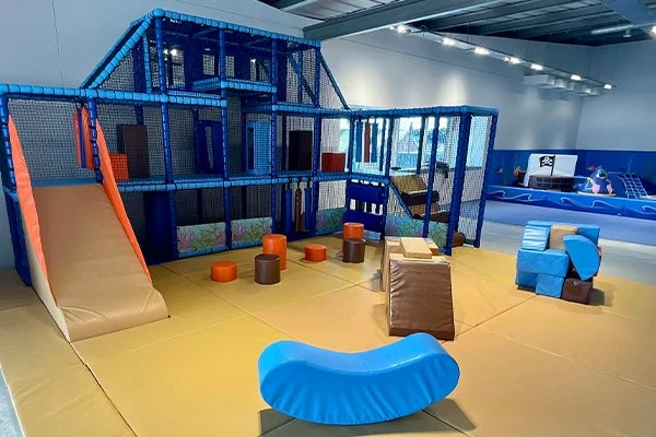 Indoor children's play area with a climbing structure, slide, padded stepping stones, foam blocks, and a soft obstacle course on yellow mats.
