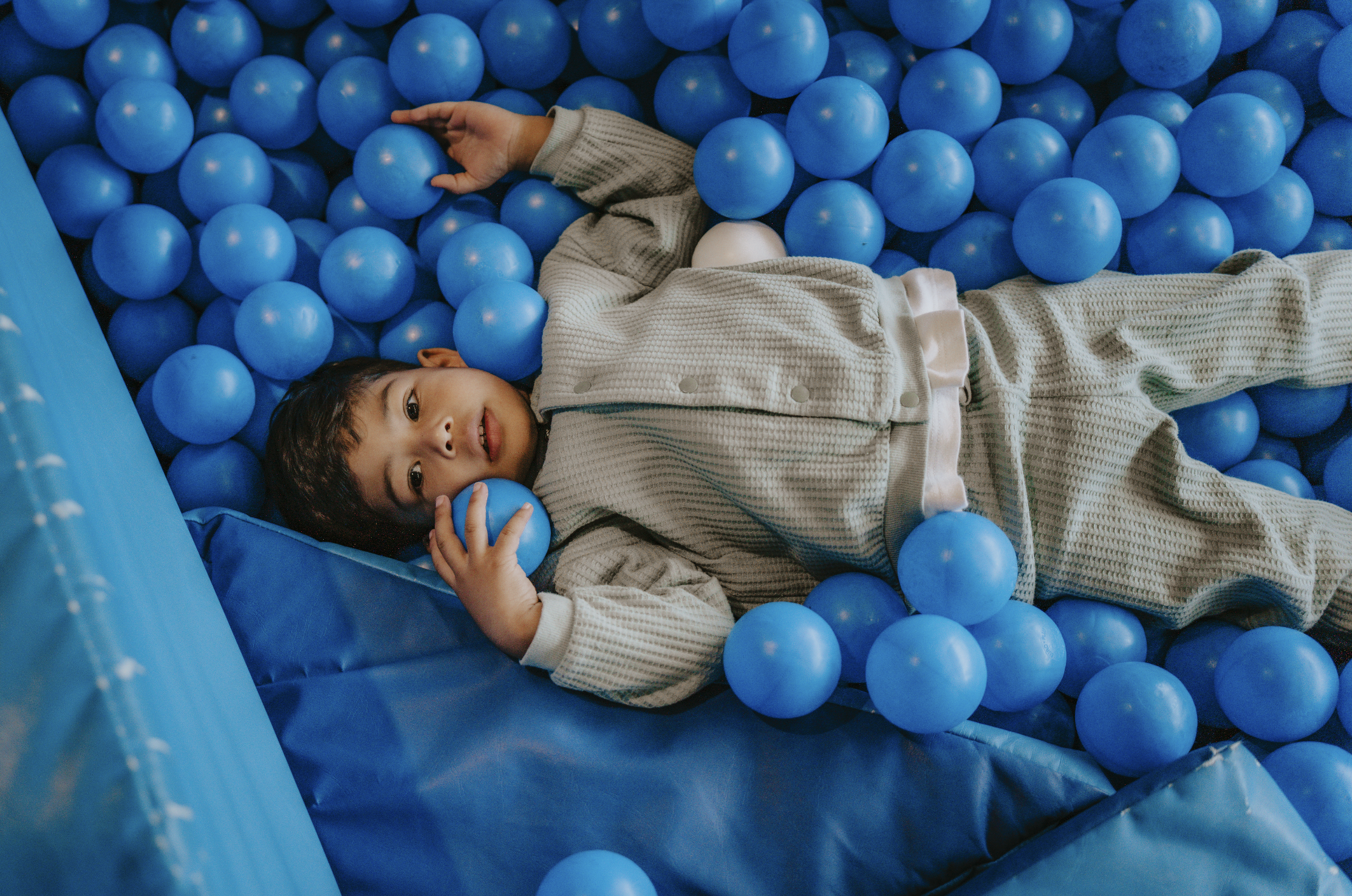 A young child lying in a ball pit filled with blue plastic balls, holding one ball with a peaceful expression.