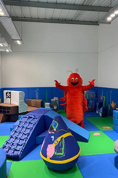 An indoor children's play area featuring a person in a red lobster costume with a smiling face, arms outstretched, surrounded by colorful soft play equipment and walls decorated with aquatic themes.