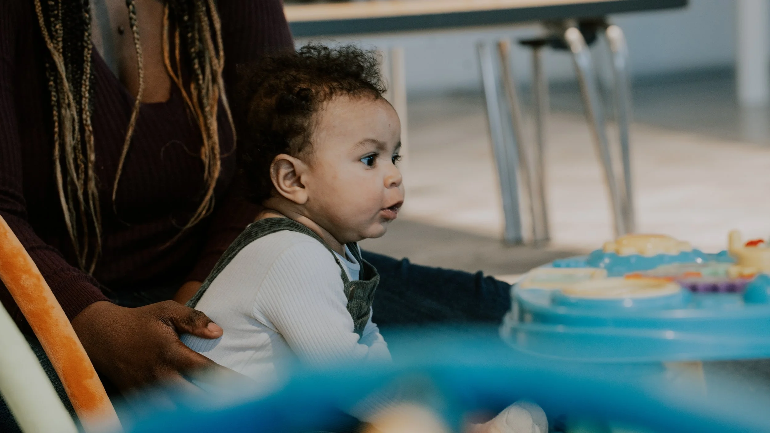 A young child with curly hair sitting at a table, looking intently at colorful toys or activities in front of him, while an adult supports him with a hand on his back. The scene appears to be in a bright, indoor setting.