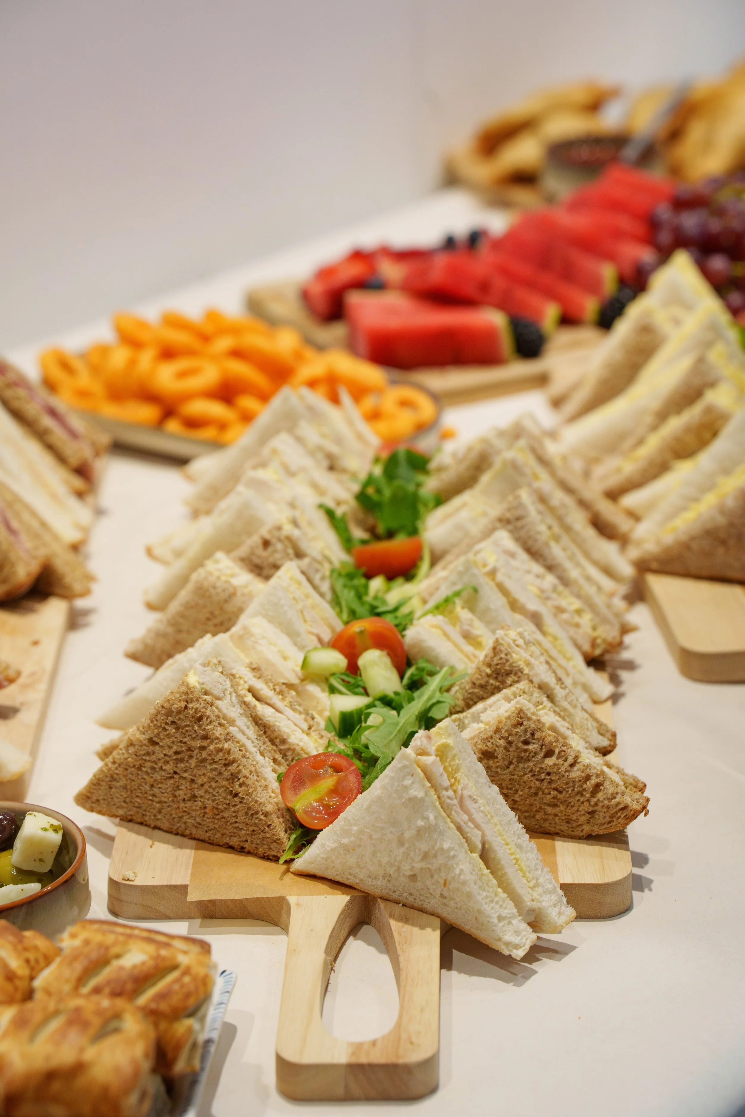 Assorted finger sandwiches with vegetables, surrounded by trays of fruit and other snacks on a white table.