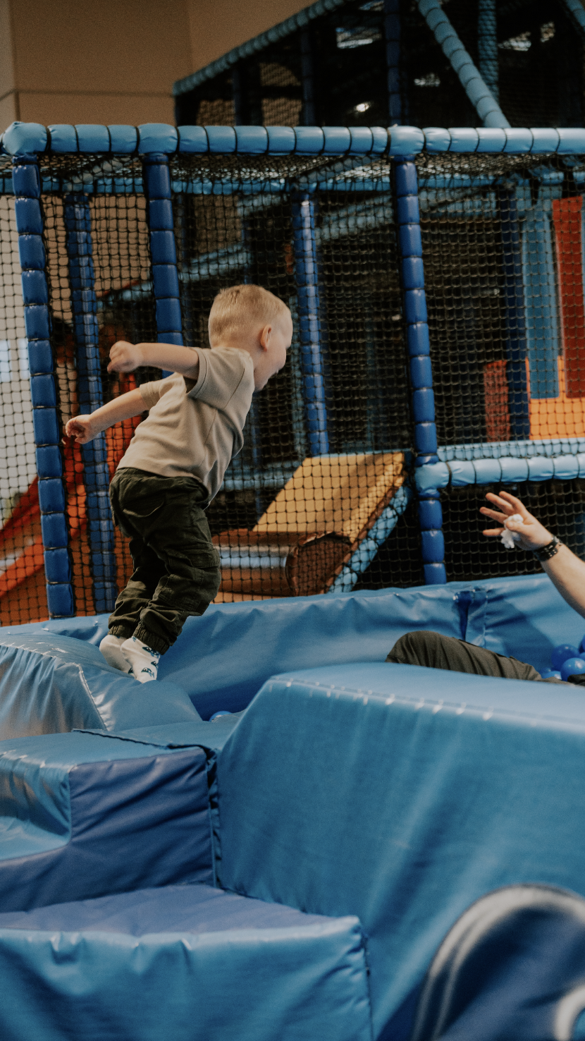 A young boy with blond hair is jumping on an indoor trampoline surrounded by safety padding and netting at an indoor playground.