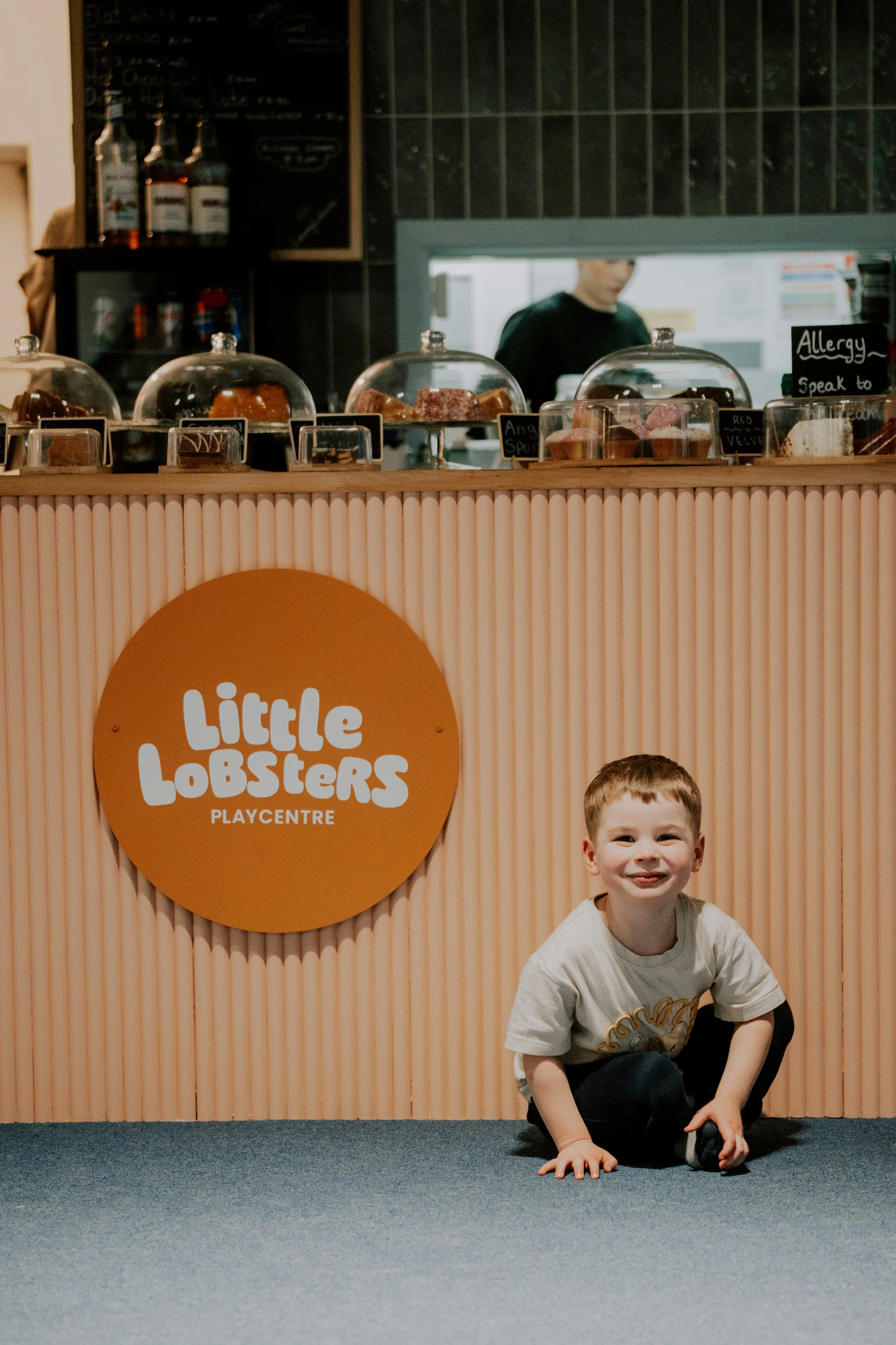 A boy with red hair and a white T-shirt crouches on blue carpet in front of a Little Lobsters Playcentre sign, smiling at the camera. Behind him, a counter with desserts under glass covers and a black chalkboard sign is visible, along with a person i