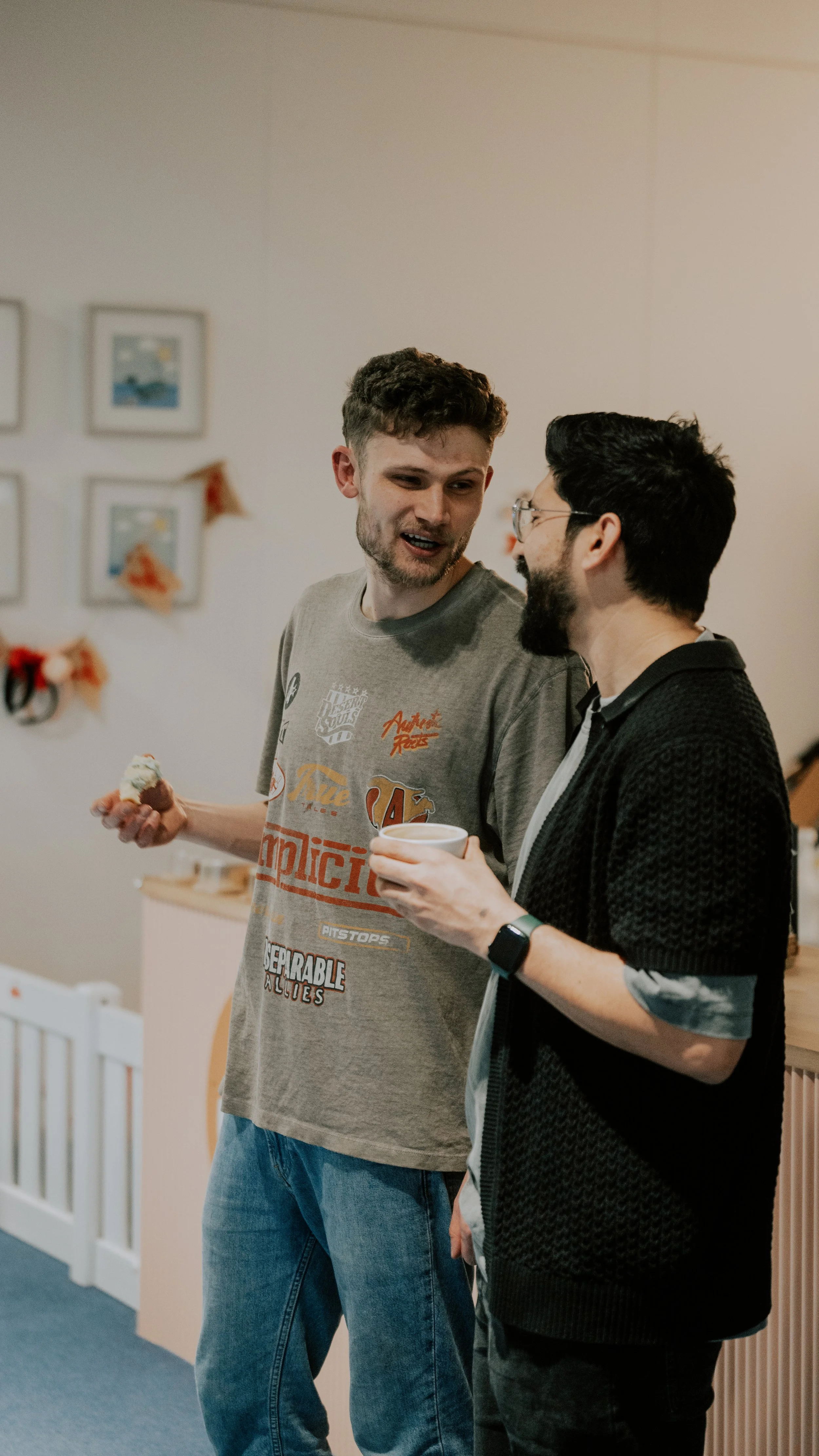 Two men are standing and talking indoors, one holding a cupcake and the other holding a cup, in a room decorated with small framed pictures and festive ornaments.