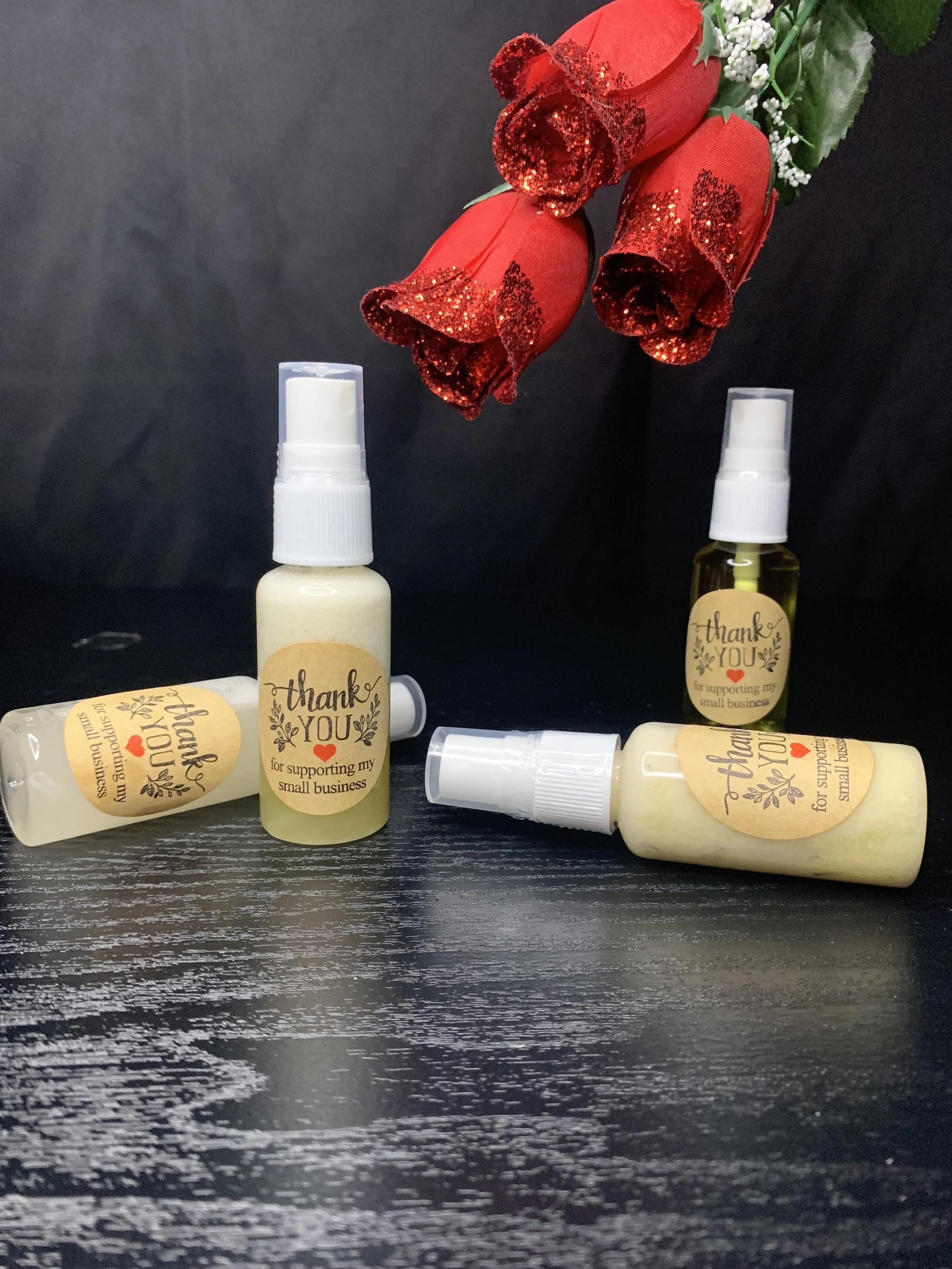 Three small spray bottles with a thank you label for supporting a small business, placed on a dark surface with red decorative flowers in the background.