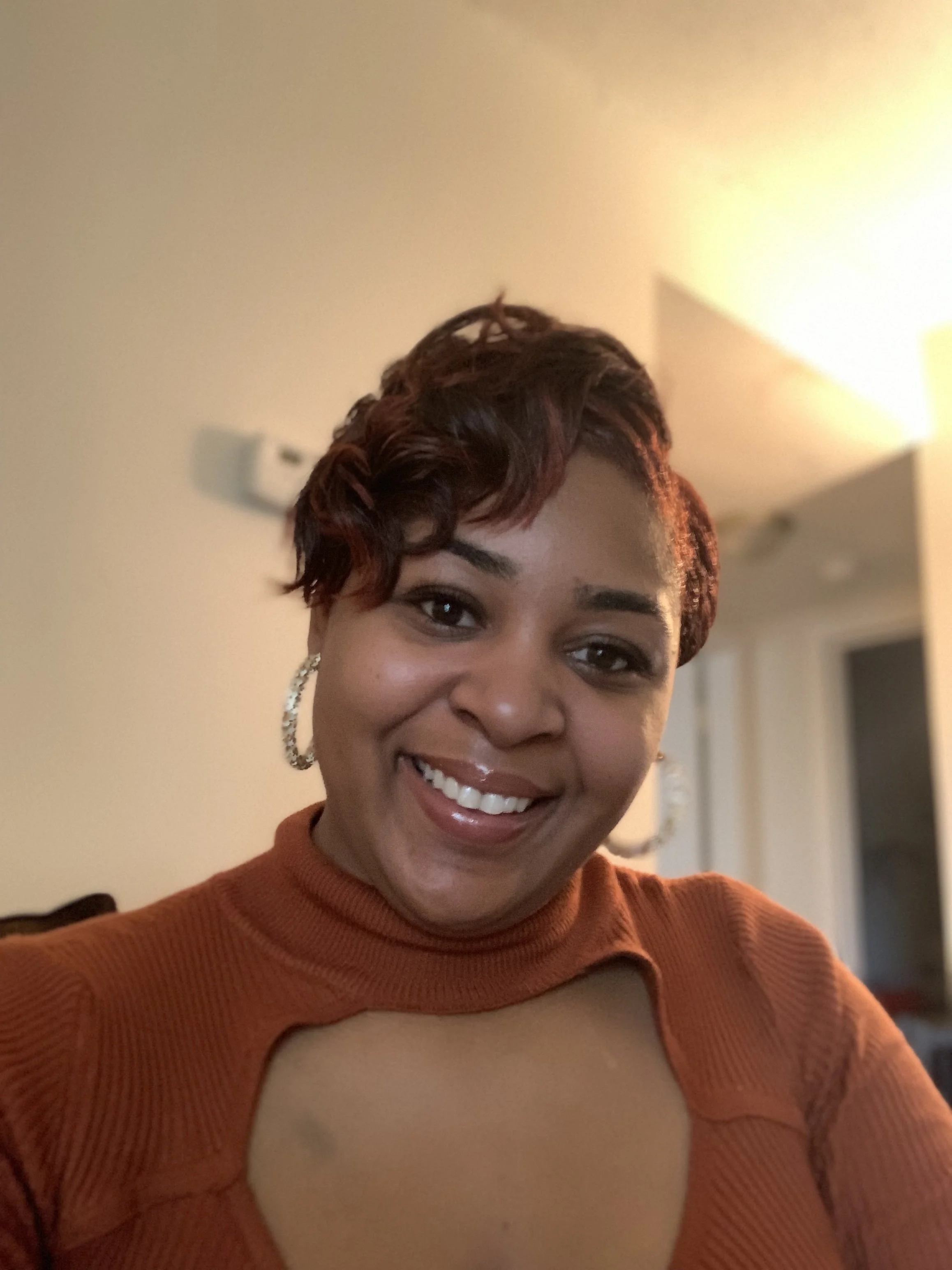 A smiling woman with short, wavy, reddish-brown hair, wearing hoop earrings and an orange top with a keyhole cutout.