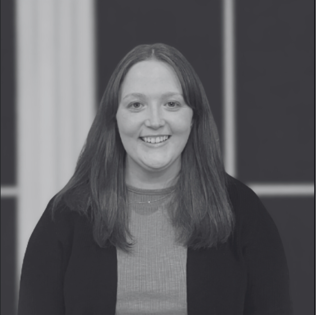 Black and white portrait of a woman with long, straight hair, smiling, wearing a dark cardigan over a light-colored top, with a blurred background that includes vertical elements.