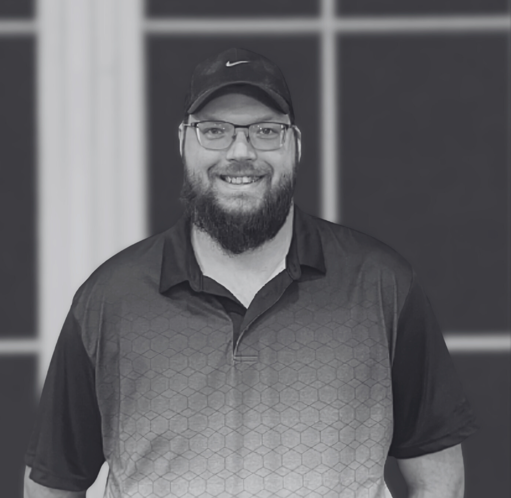 A man with a beard, glasses, and a black cap smiling at the camera. He is wearing a polo shirt with a hexagonal pattern on the front. The background features large windows or glass panels.