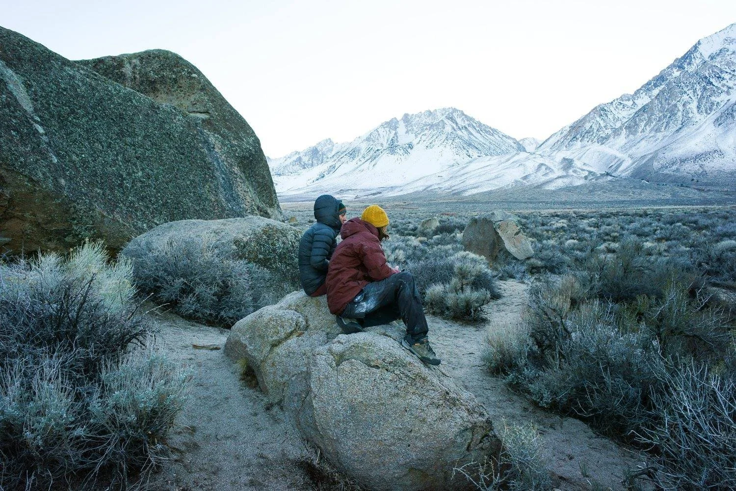 Two people sitting on a large rock in a snowy mountainous landscape with sparse vegetation and snow-covered peaks in the background.